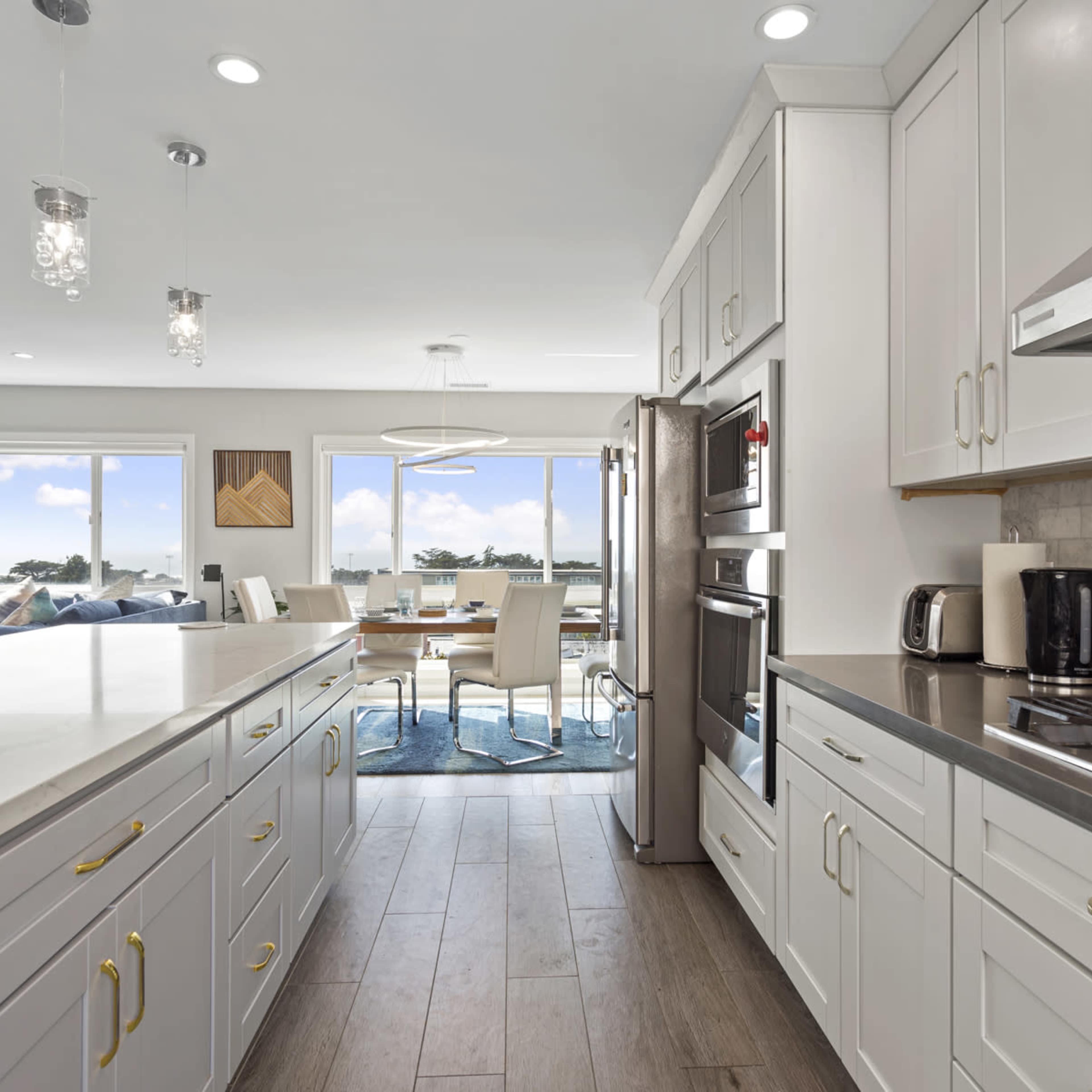 A modern kitchen features white cabinetry, a large island with a marble countertop, and stainless steel appliances, overlooking a dining area with large windows revealing a view of the outdoors.