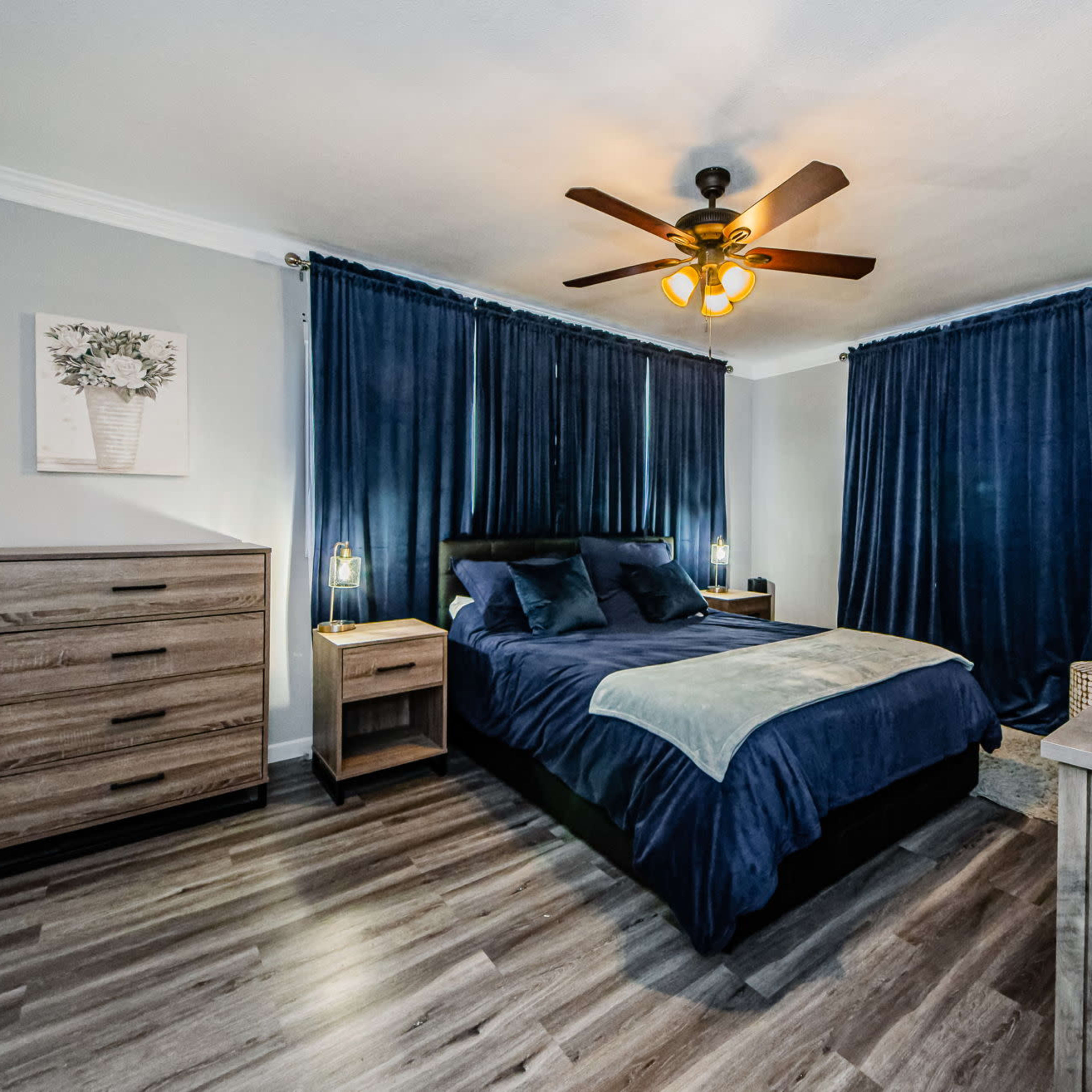 A modern bedroom features a large bed with dark bedding, wooden furniture, and blue curtains, illuminated by a ceiling fan with a light fixture.