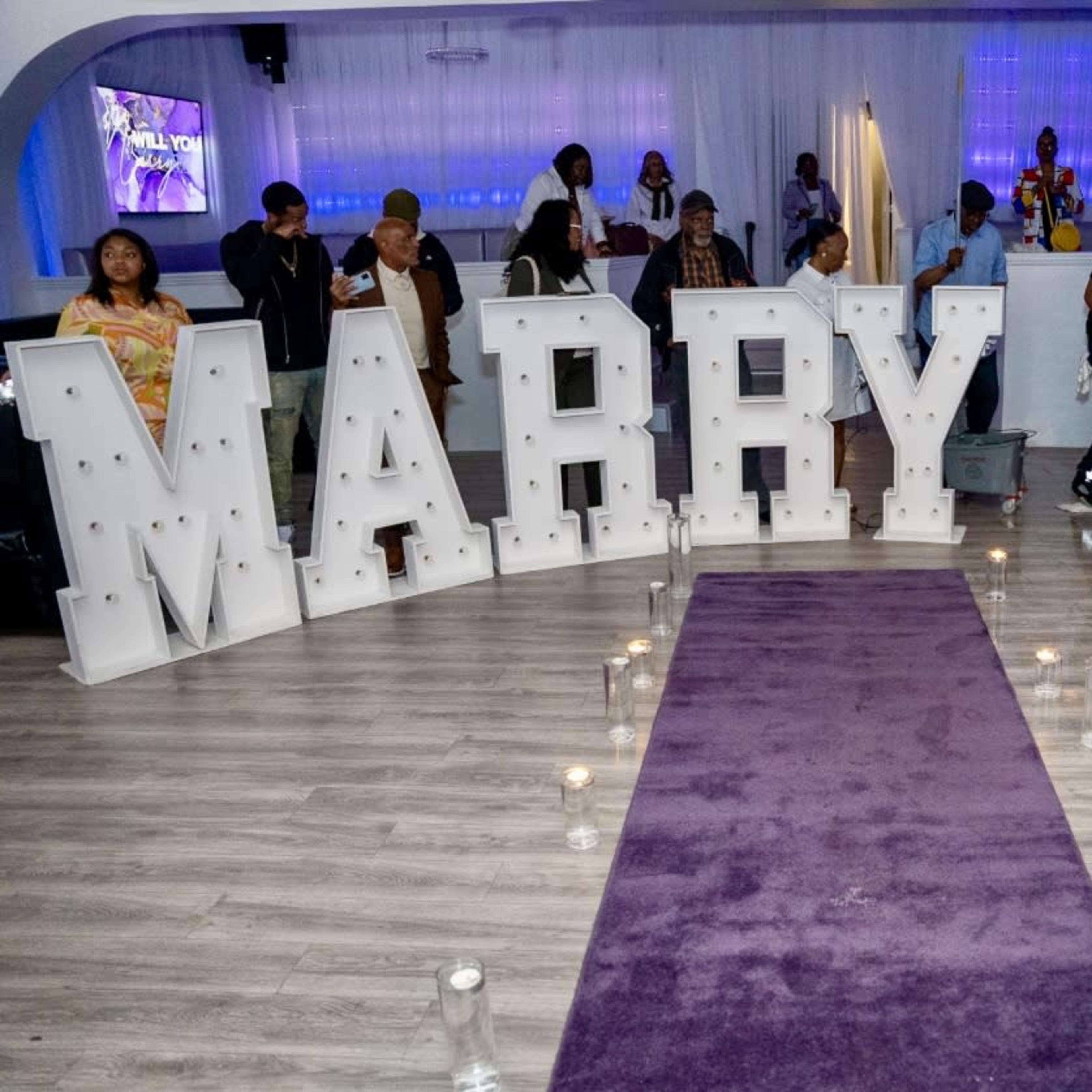 A large decorative display spelling "MARRY ME" is set up in a venue with guests mingling around a purple carpet and candles.
