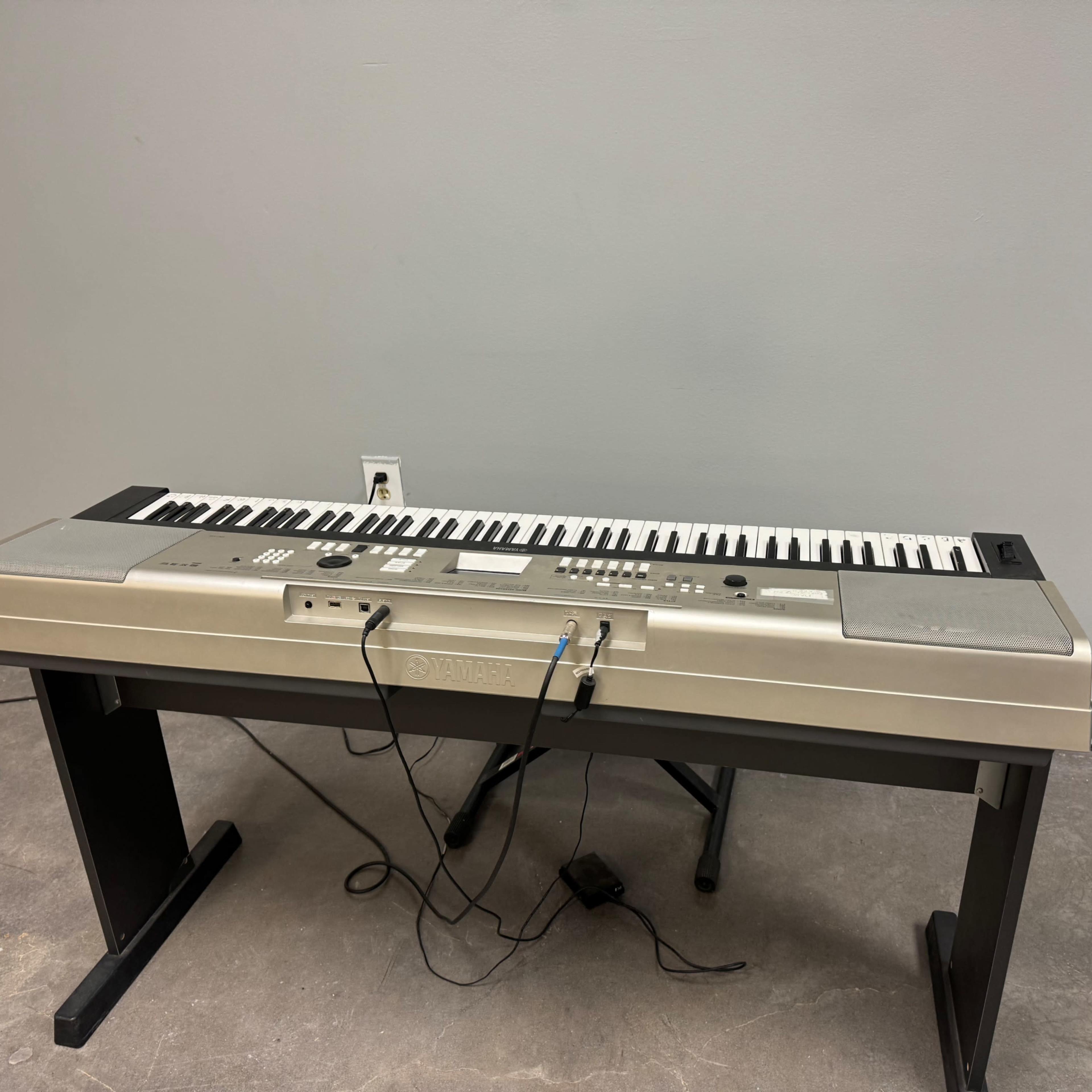A silver electronic keyboard placed on a black stand against a plain gray wall.