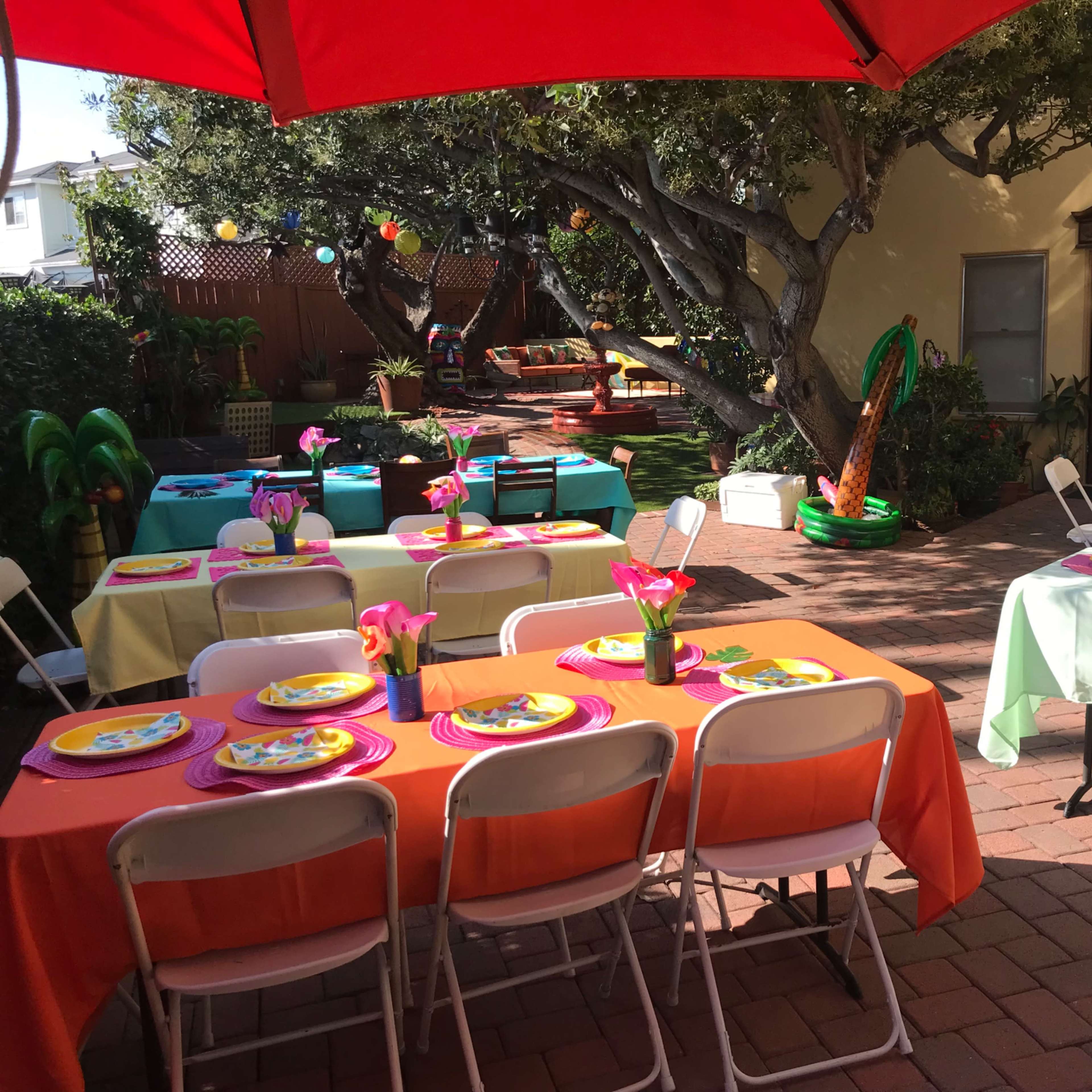 The scene features a vibrant outdoor party setup with decorated tables under umbrellas, set amidst lush greenery.