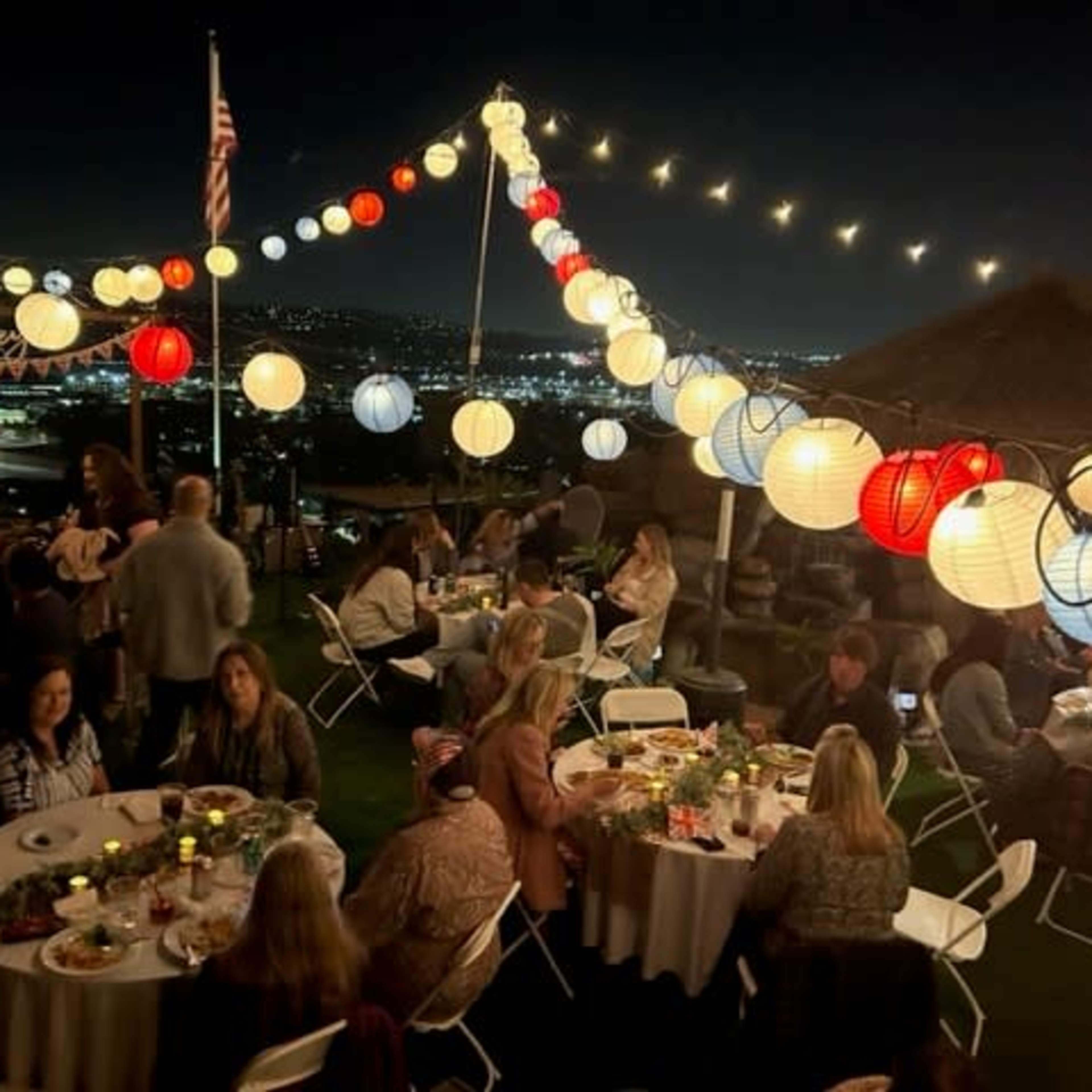 A group of people gathers around tables adorned with food and drinks, illuminated by colorful lanterns and string lights against a night sky overlooking a cityscape.