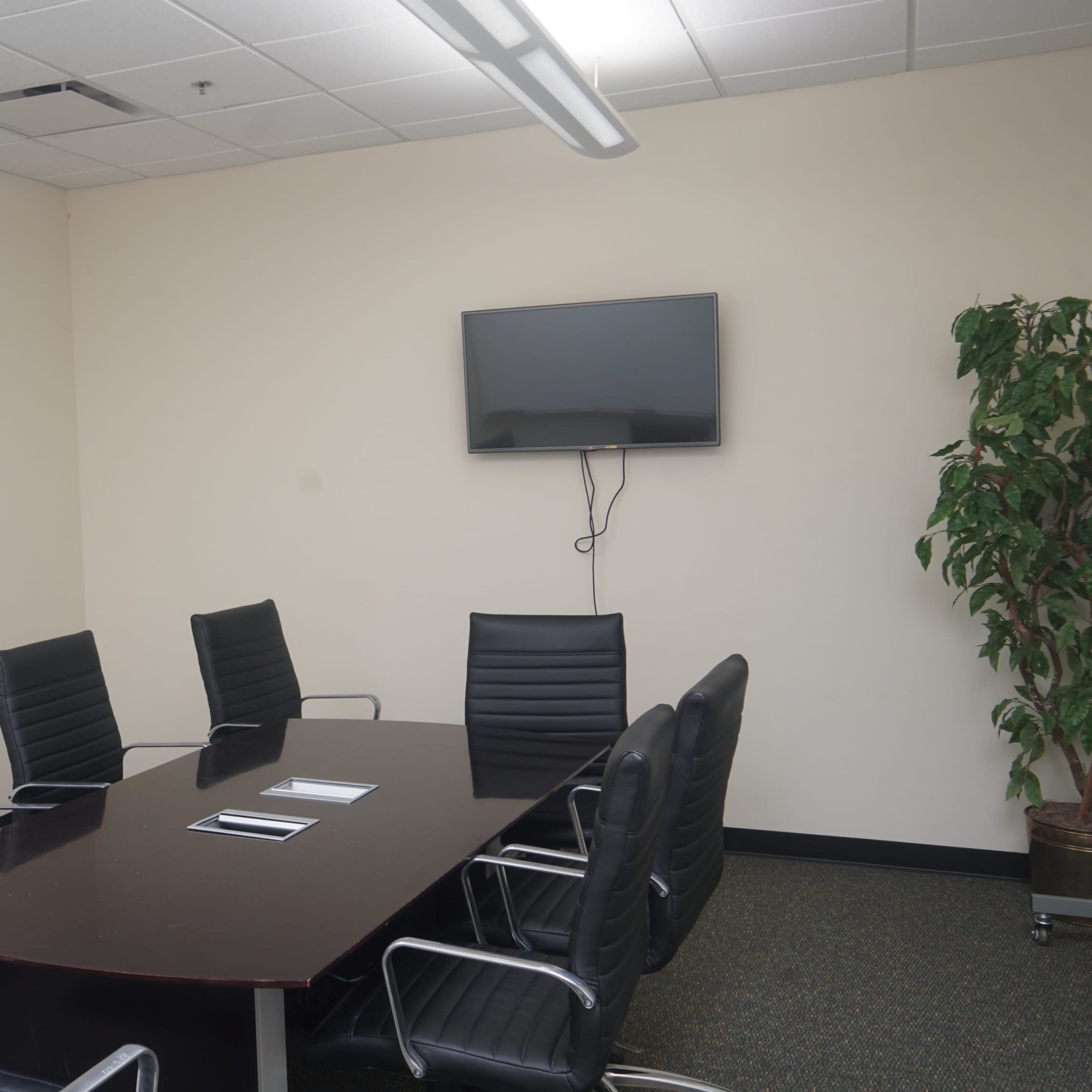 A conference room features a large rectangular table surrounded by black leather chairs, with a television mounted on the wall and a glass whiteboard.