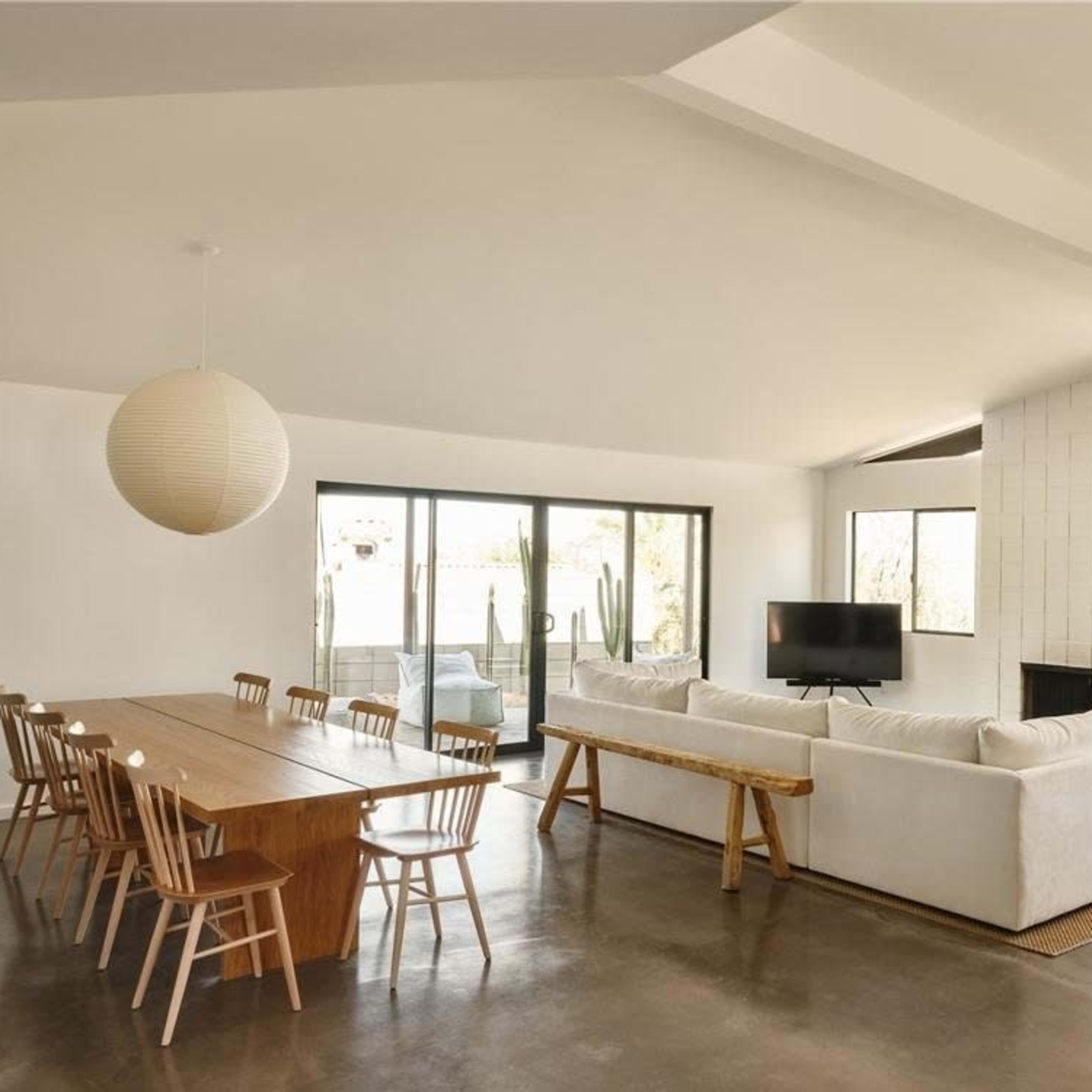 A modern living space featuring a dining table with chairs, a sectional sofa, a television, and large windows with views of greenery.