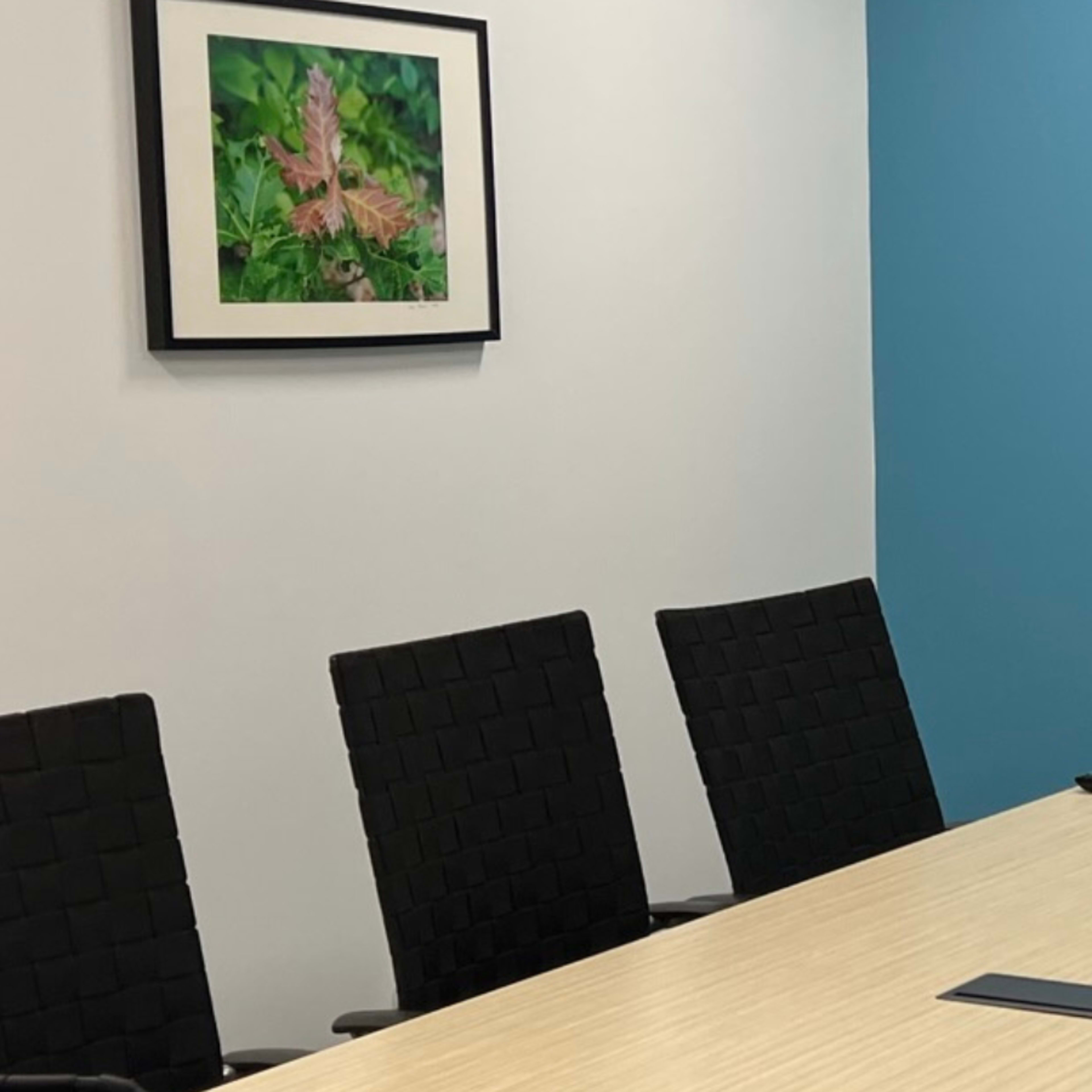 A conference room features a wooden table surrounded by six black chairs, with framed artwork on the walls and a television mounted on a blue wall.