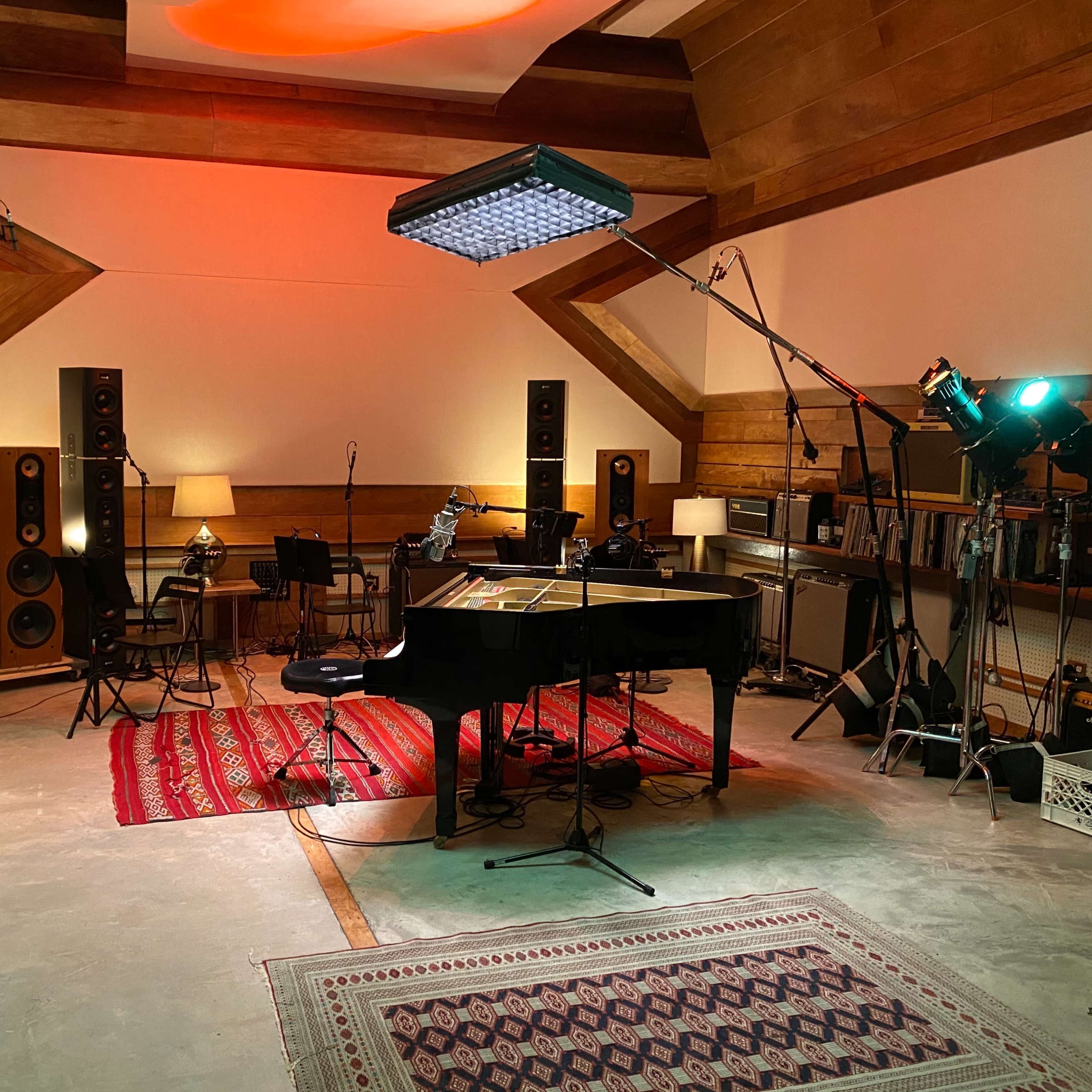 A modern recording studio features a grand piano, multiple speakers, and various lighting equipment arranged around a colorful area rug.