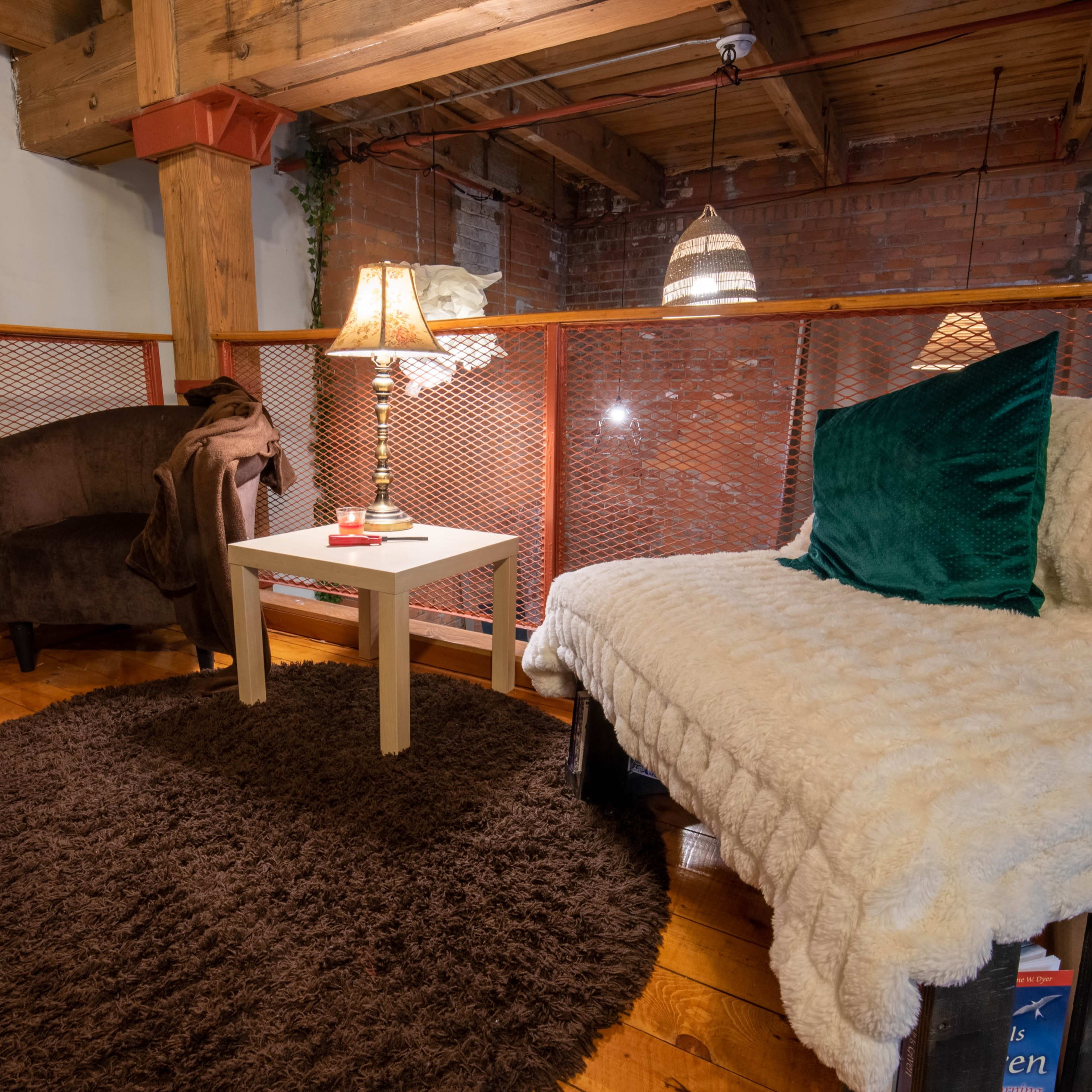 A cozy living room corner features a plush sofa and a chair beside a small table with a lamp, surrounded by wooden flooring and exposed brick walls.