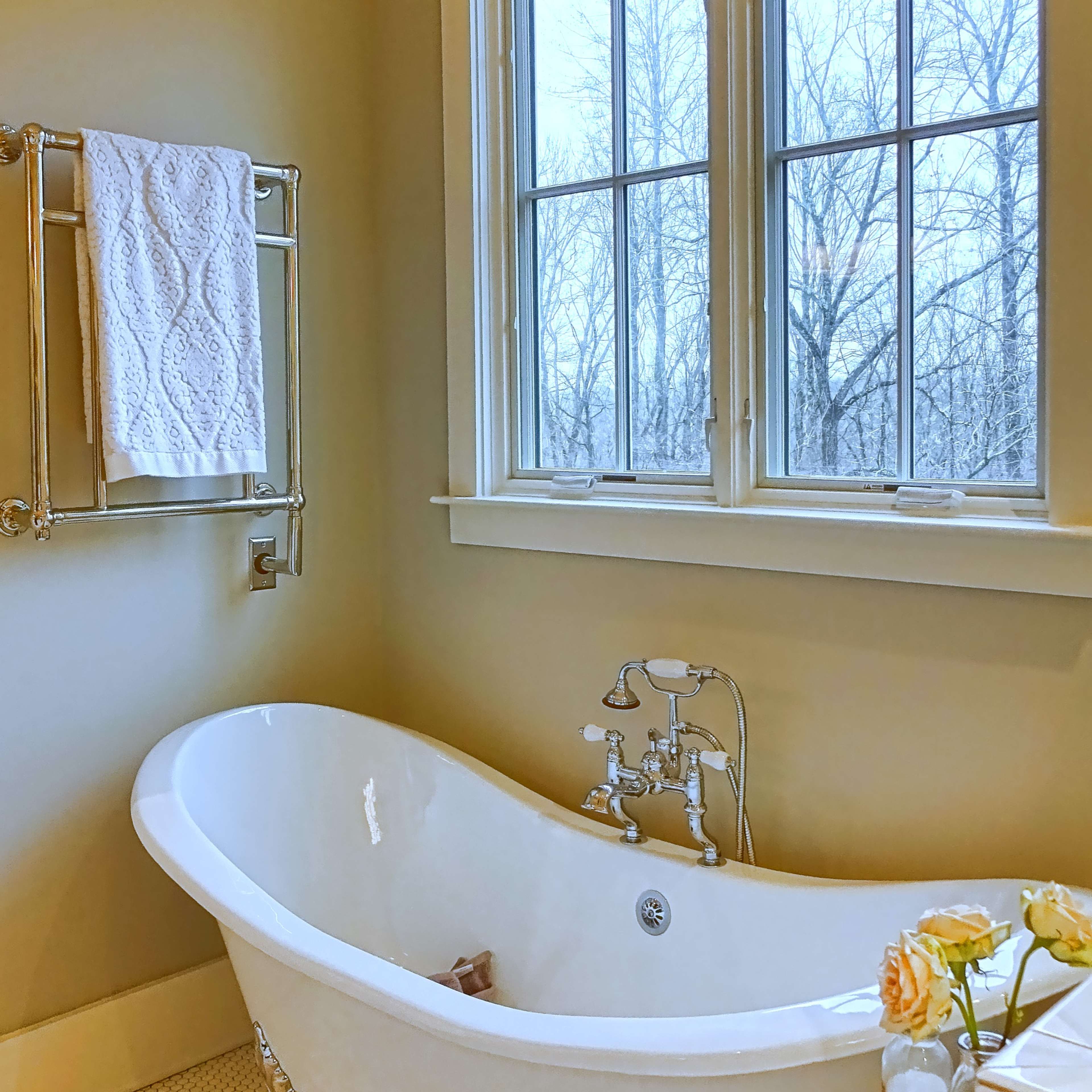 A white freestanding bathtub is positioned next to a window in a bathroom with light-colored walls and a tile floor.