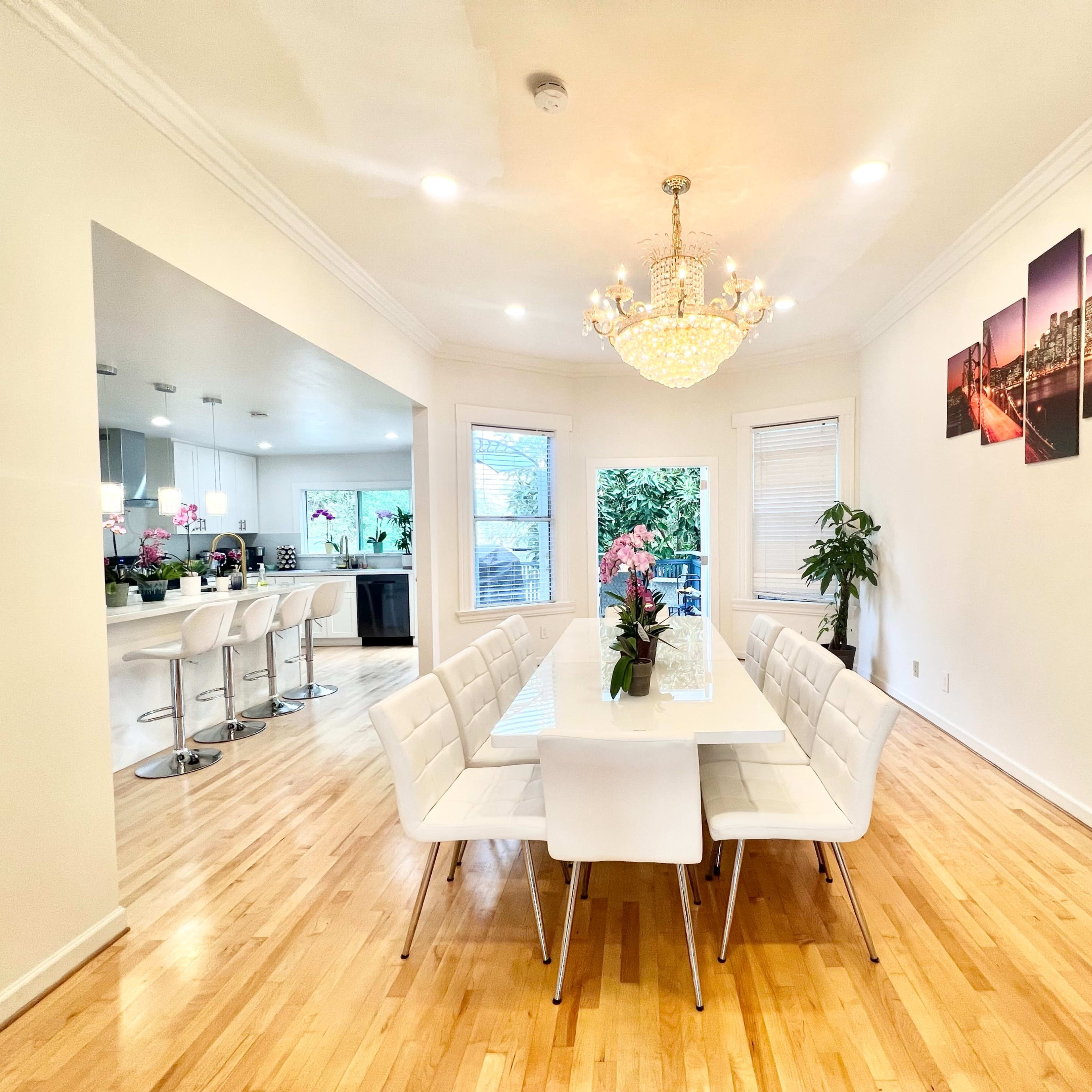 A modern dining room features a long white table surrounded by white chairs, with a chandelier above and a large window revealing kitchen views.