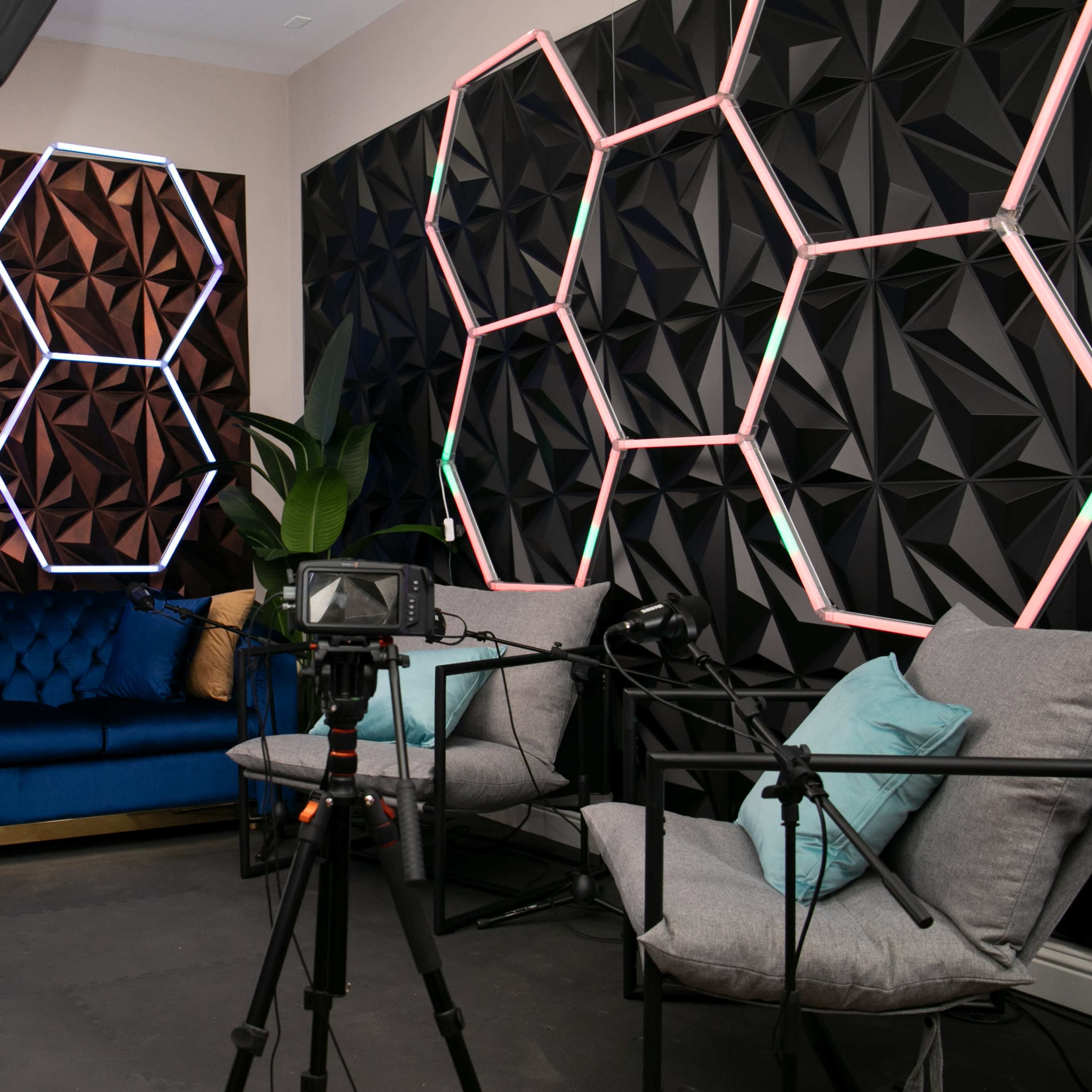A modern interior studio setup with a blue sofa, geometric-patterned wall panels, and two gray chairs, accompanied by camera equipment and lighting.