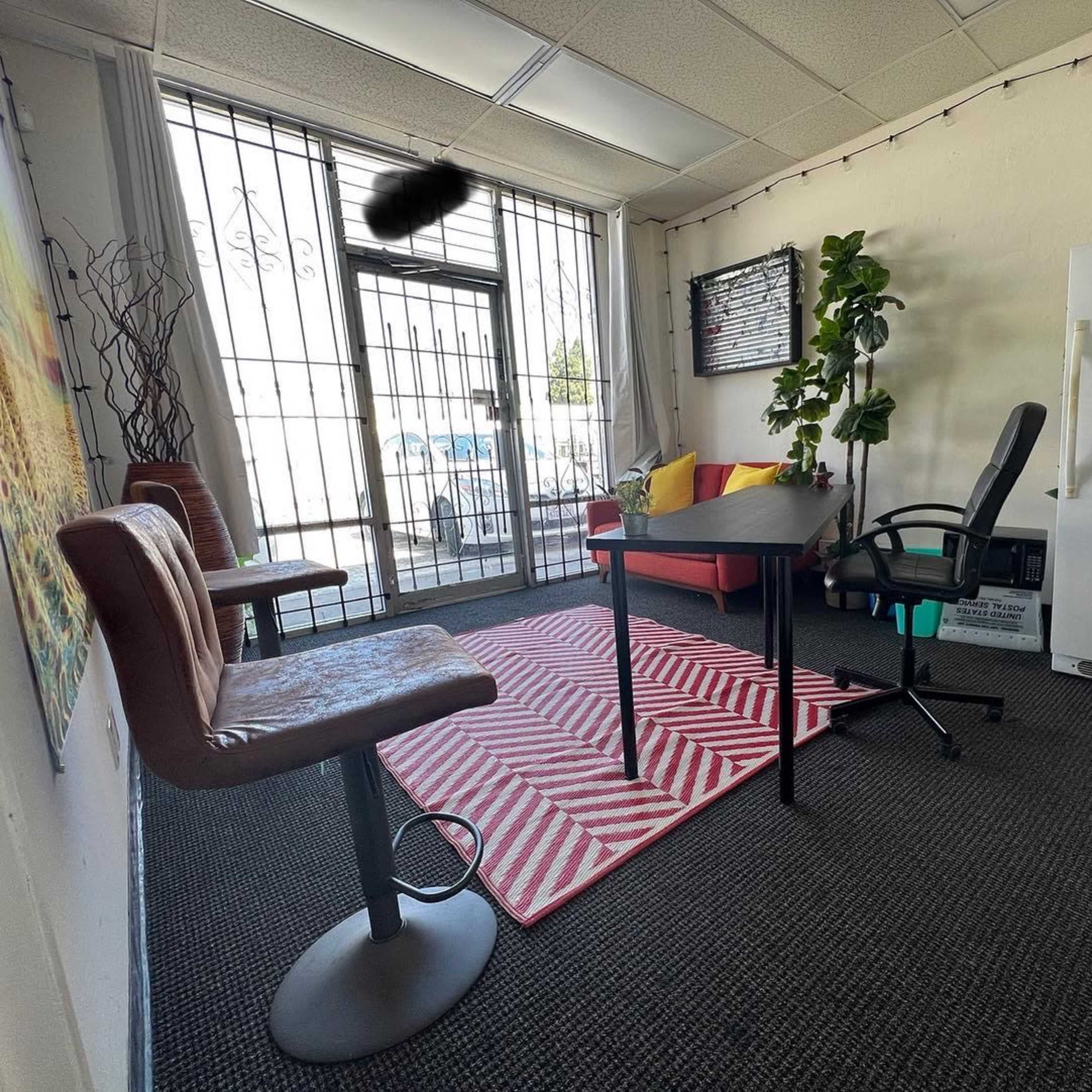 A small office space featuring a red sofa, a desk and chair, a small refrigerator, and a rug with a striped pattern, with large windows that are partially barred.