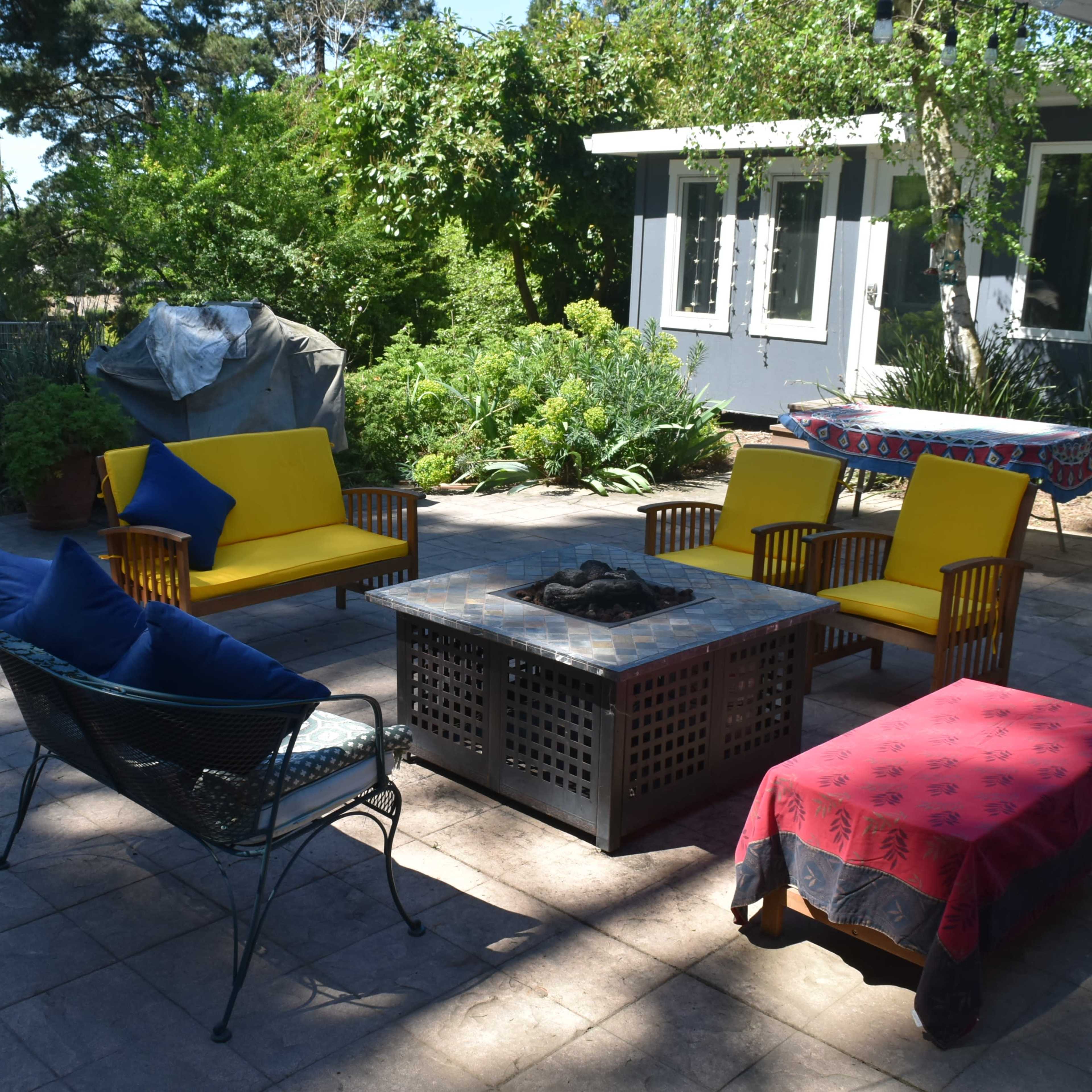 A patio features a rectangular fire pit surrounded by yellow and blue seating, with greenery and a house in the background.