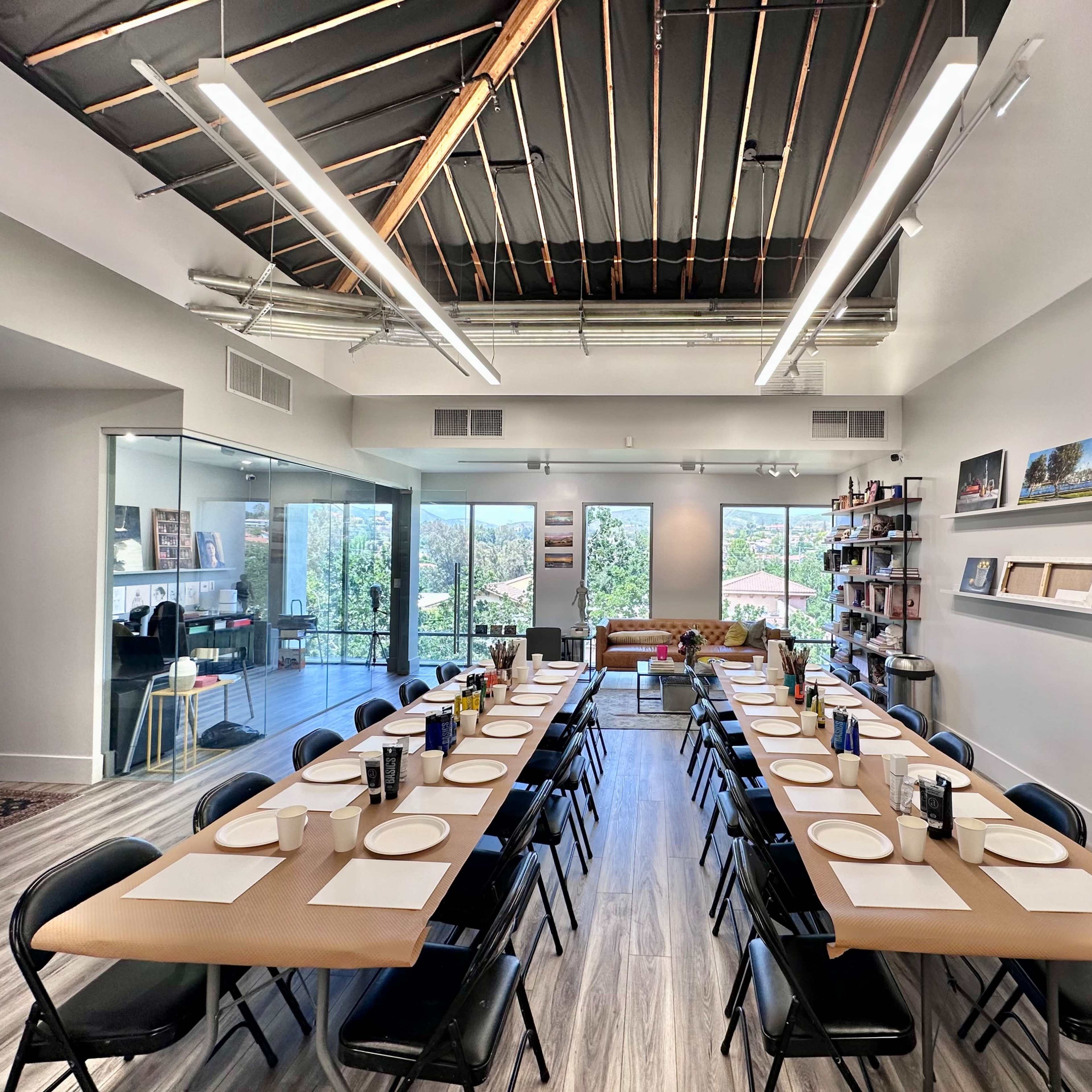 A modern dining room features a long table set with plates and utensils, flanked by black chairs, and large windows showcasing a view outside.