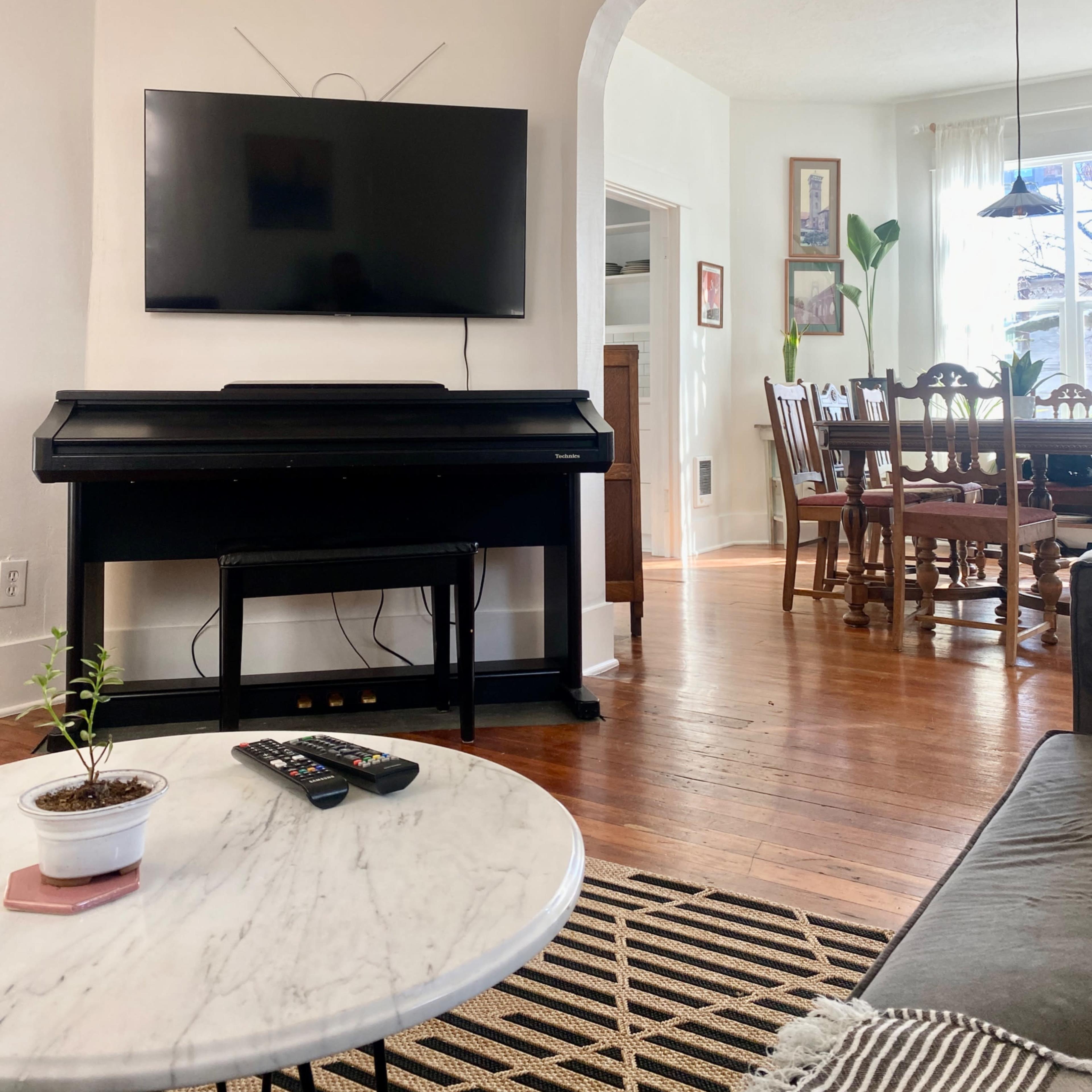 A living room with a wall-mounted television above a black piano, and a dining area with a wooden table and chairs in the background.