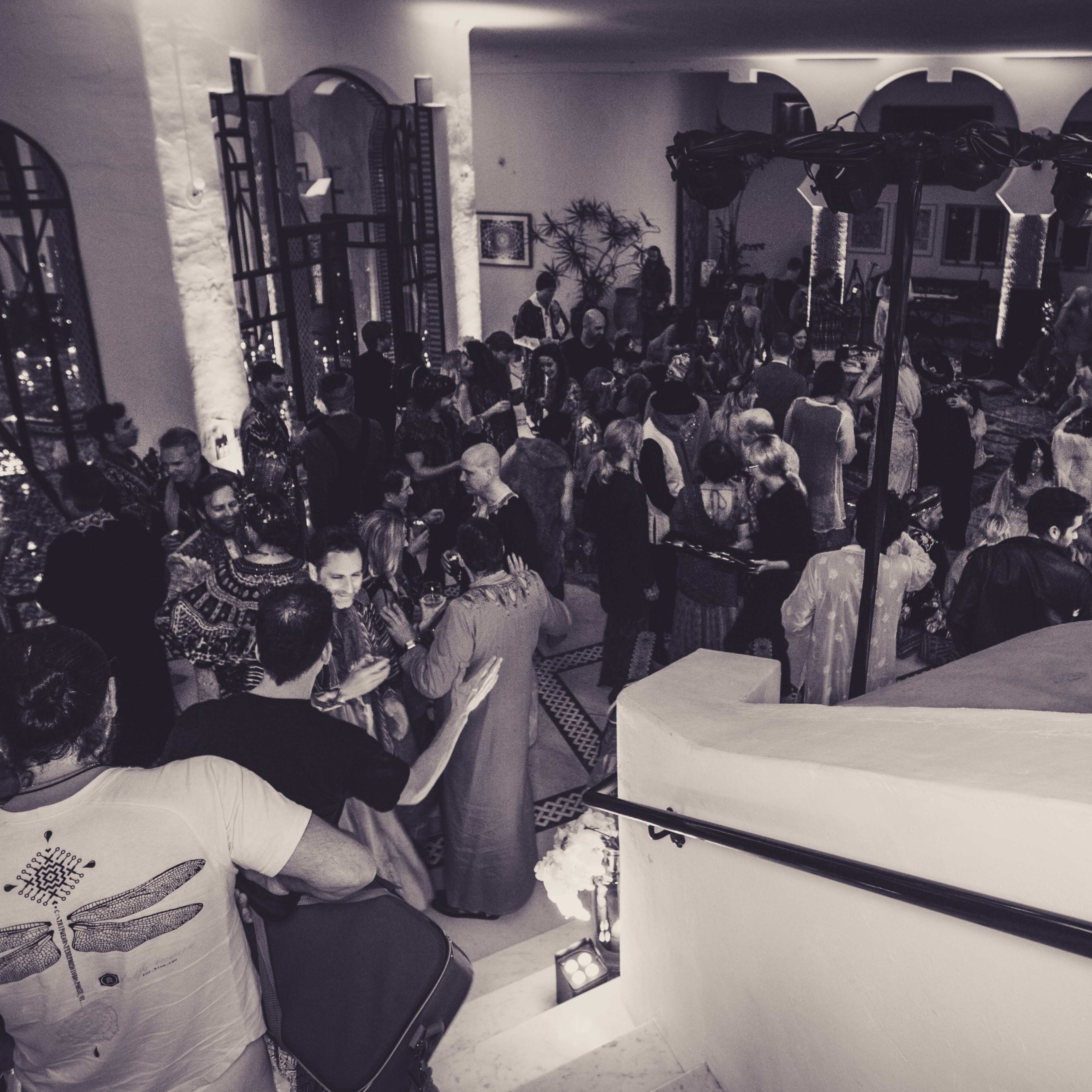 A large group of people gathers in a brightly lit hall with decorative arches and stairs, engaged in social activities.