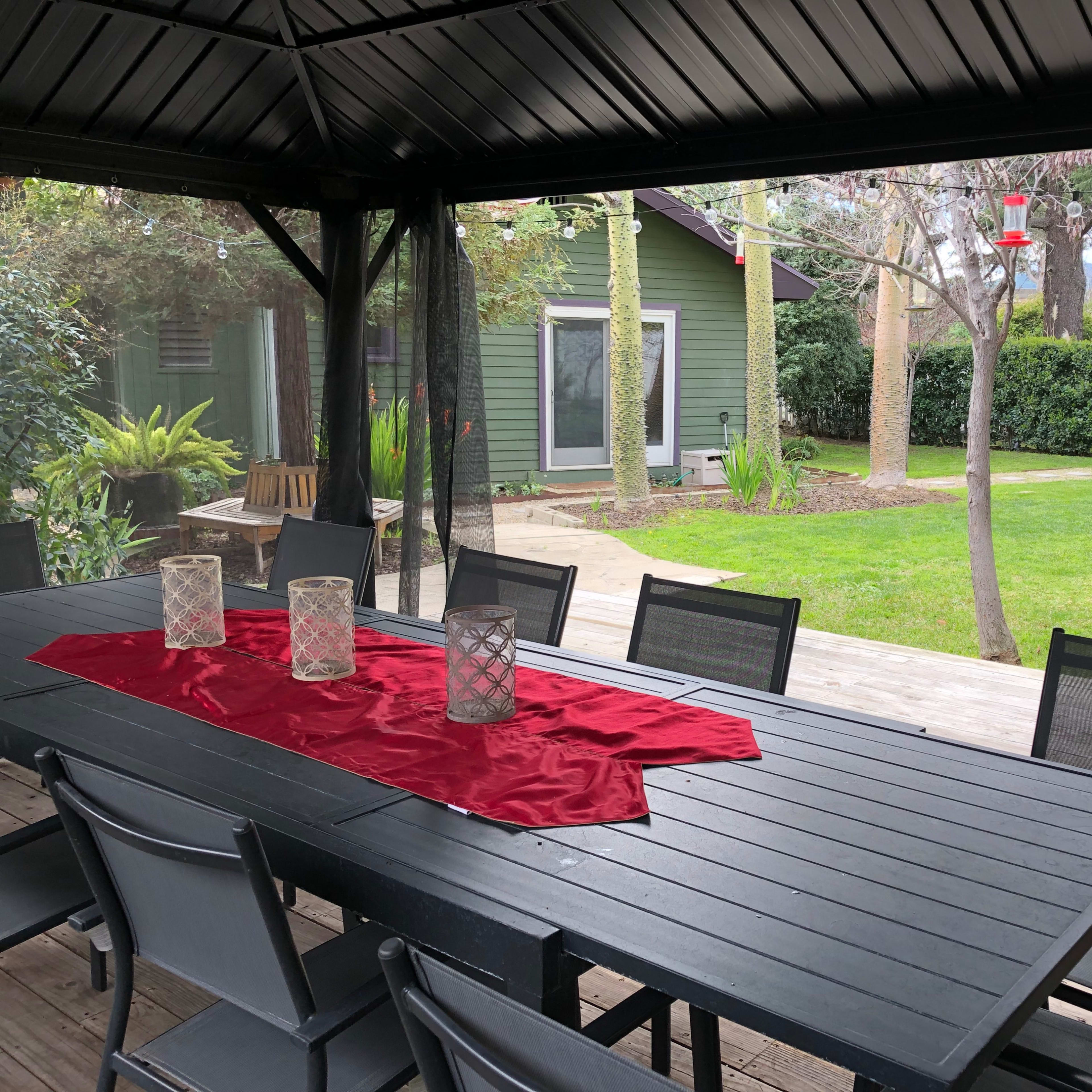 A covered outdoor dining area with a black table, surrounded by chairs, features a red table runner and candles, overlooking a green backyard with trees and shrubs.