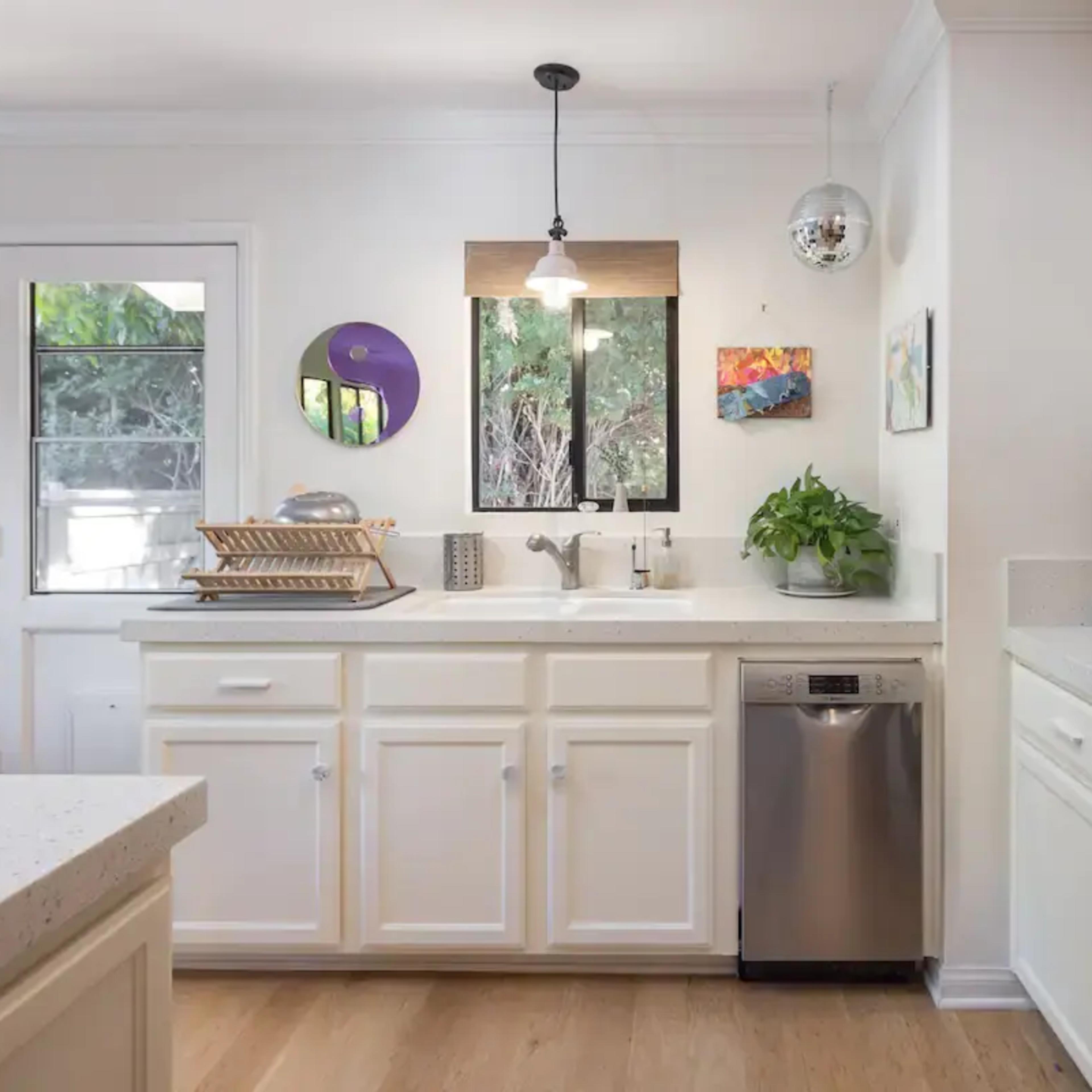 A modern kitchen features white cabinets, stainless steel appliances, and a sink beneath a window, complemented by wooden accents and decorative artwork.