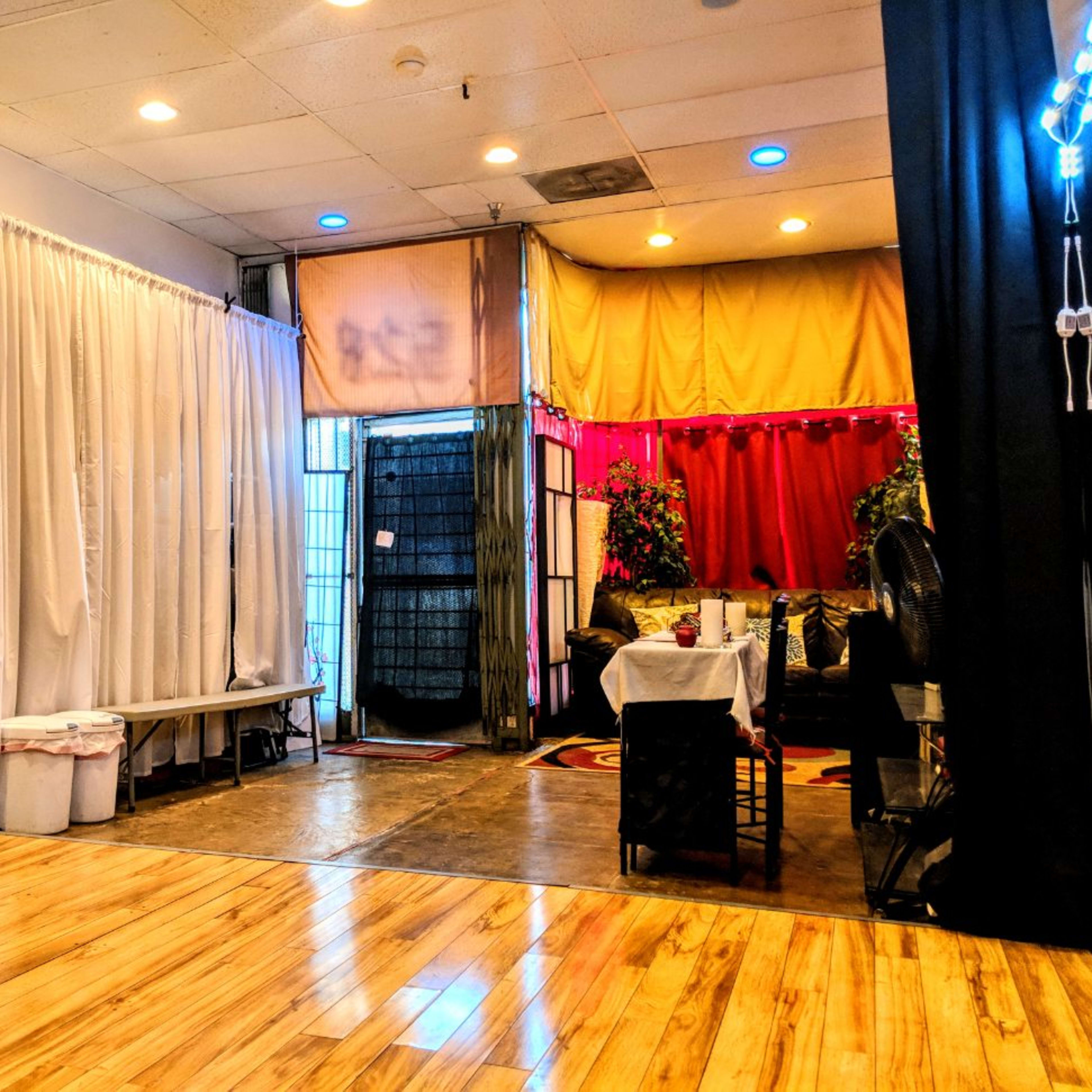 An indoor space divided by curtains, with wooden flooring, a table set up in the center, and various plants in the background.