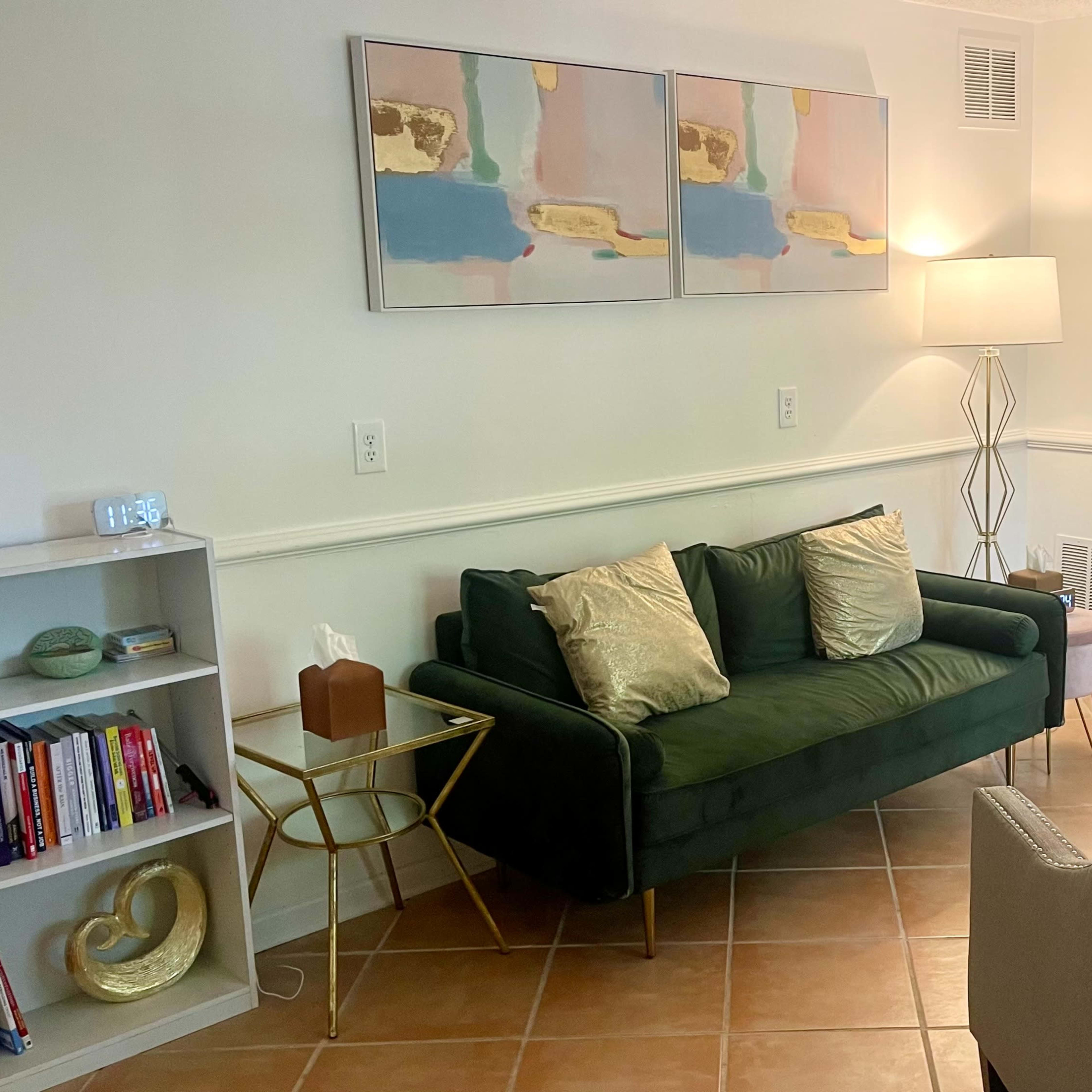 A living room features a green sofa with decorative pillows, a light-colored armchair, a small round table, a bookshelf filled with books, and abstract artwork on the wall.