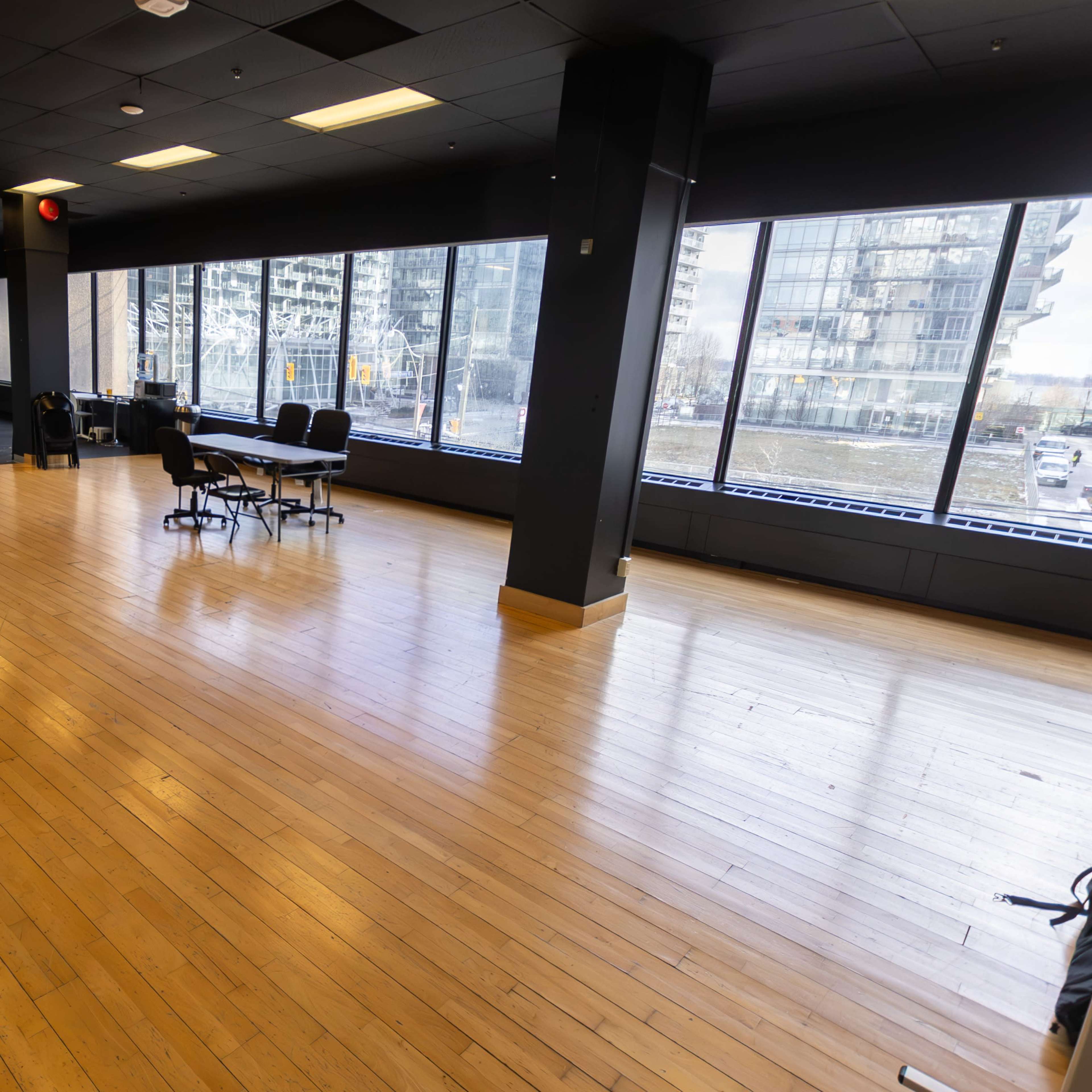 A spacious, empty room with polished wooden flooring and large windows providing natural light, furnished with a few chairs and a table.