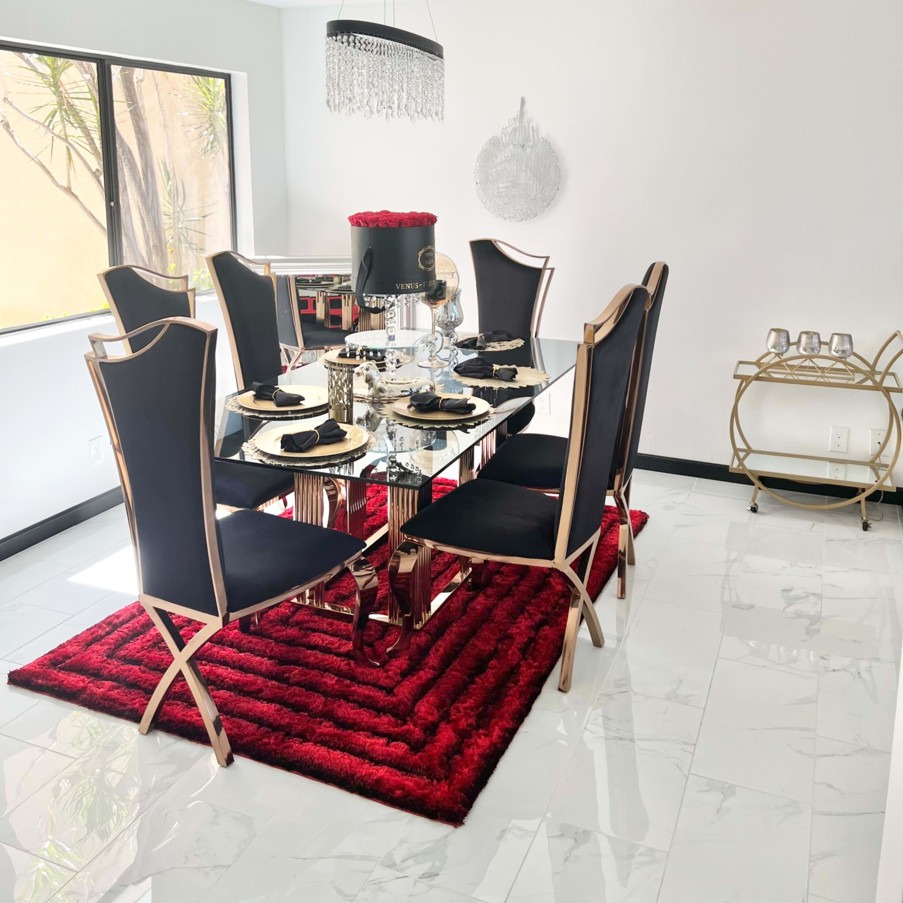 A modern dining room features a glass table surrounded by black and gold chairs, a red area rug, and a chandelier above, with a bar cart in the corner.