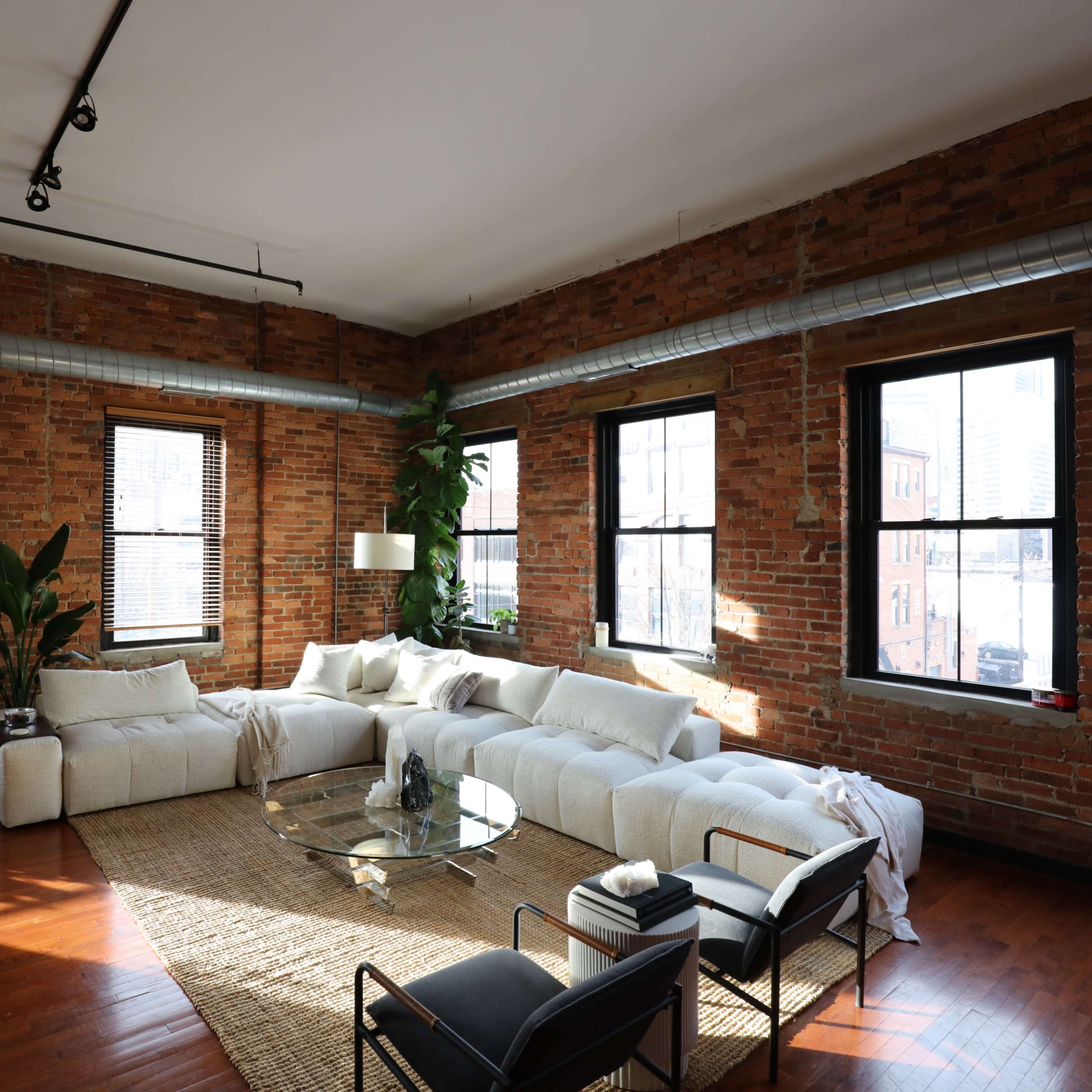 A spacious living area with exposed brick walls, large windows, a sectional sofa, and a glass coffee table on a woven rug.