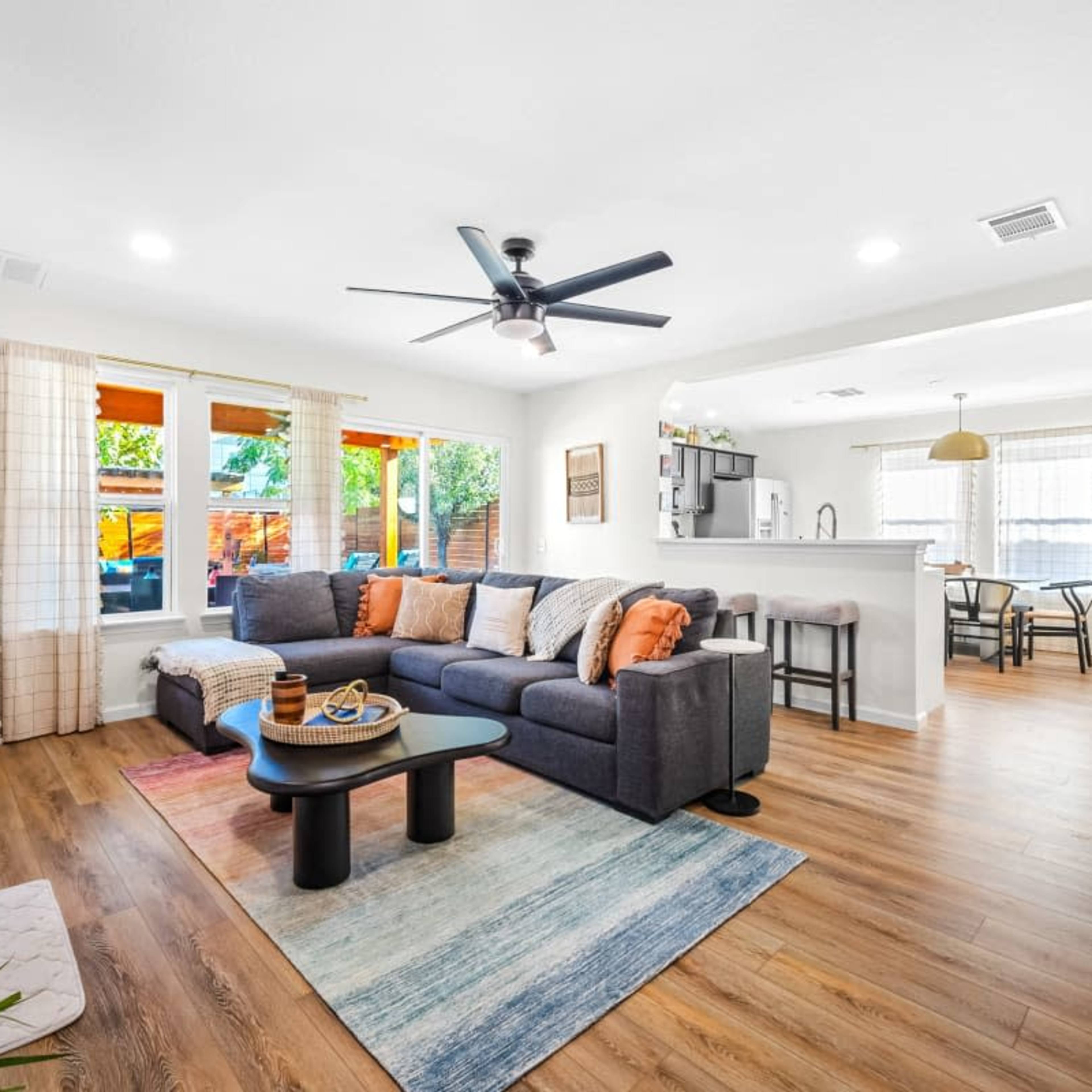 A modern living room features a large sectional sofa, a coffee table, and a television mounted on the wall, with a kitchen area and dining space visible in the background.