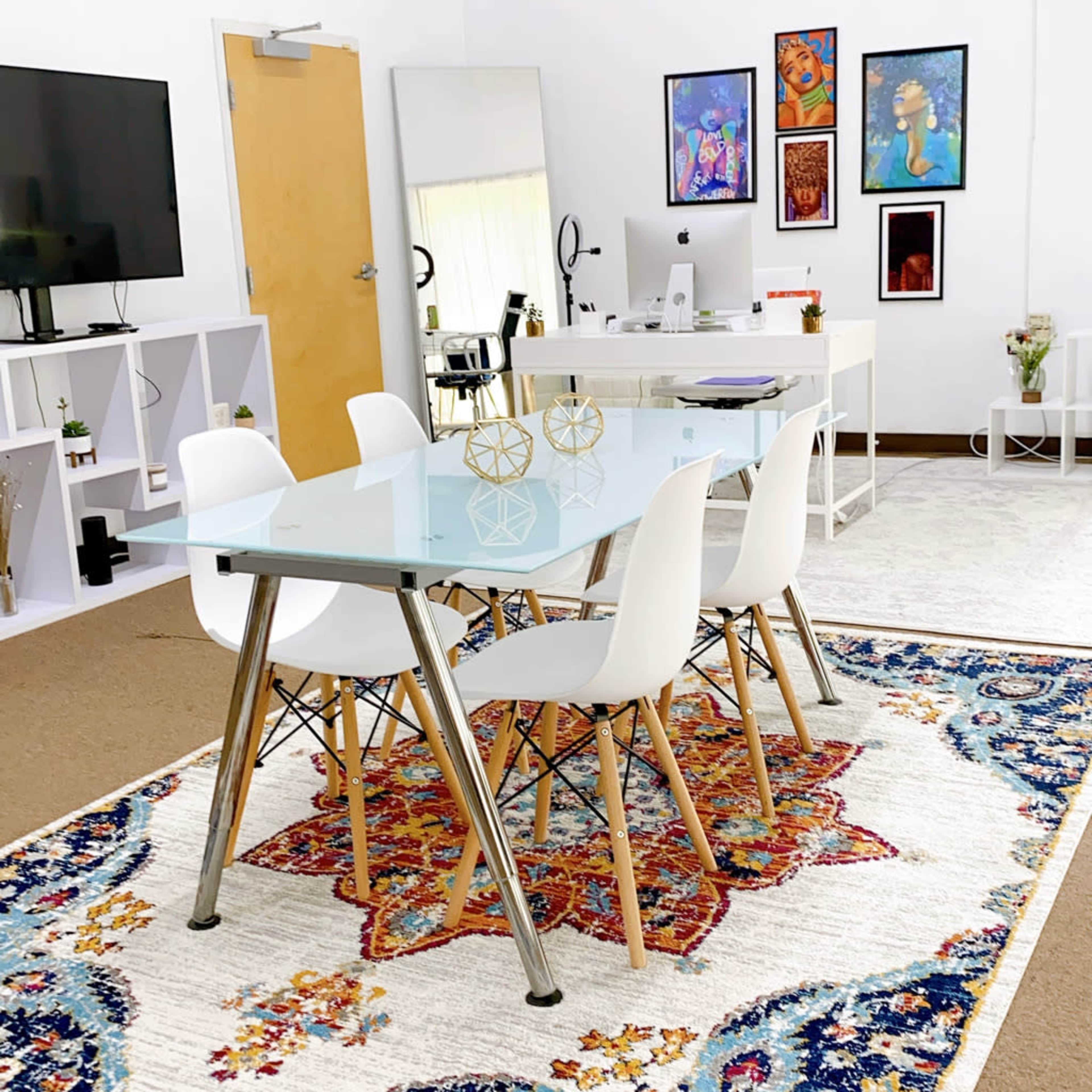 A modern office space featuring a glass-top dining table surrounded by four white chairs on a patterned rug, with a television, a desk, and various artworks on the walls.