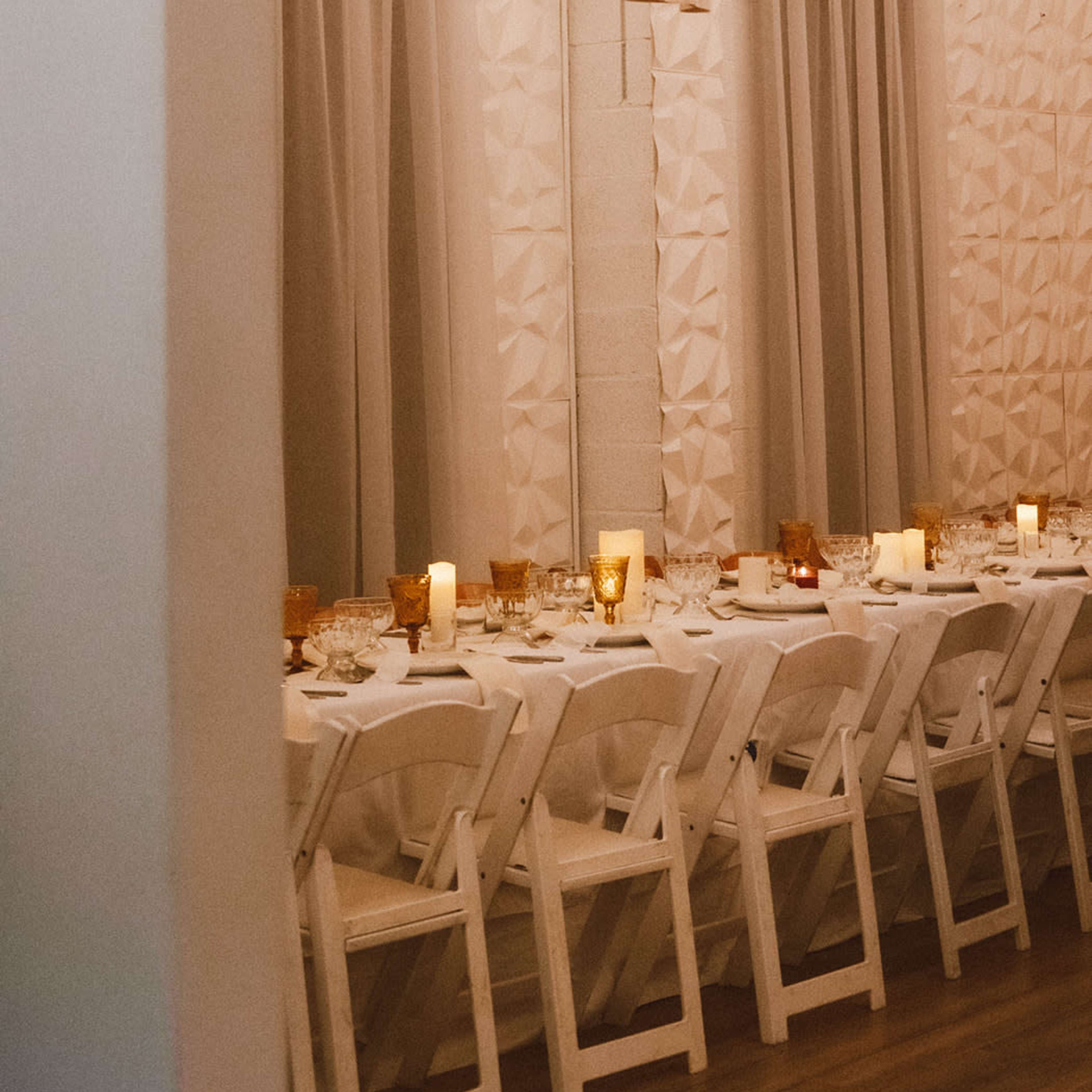 A decorated dining area features a long white table set with glassware and amber candles under soft draping and warm lighting.