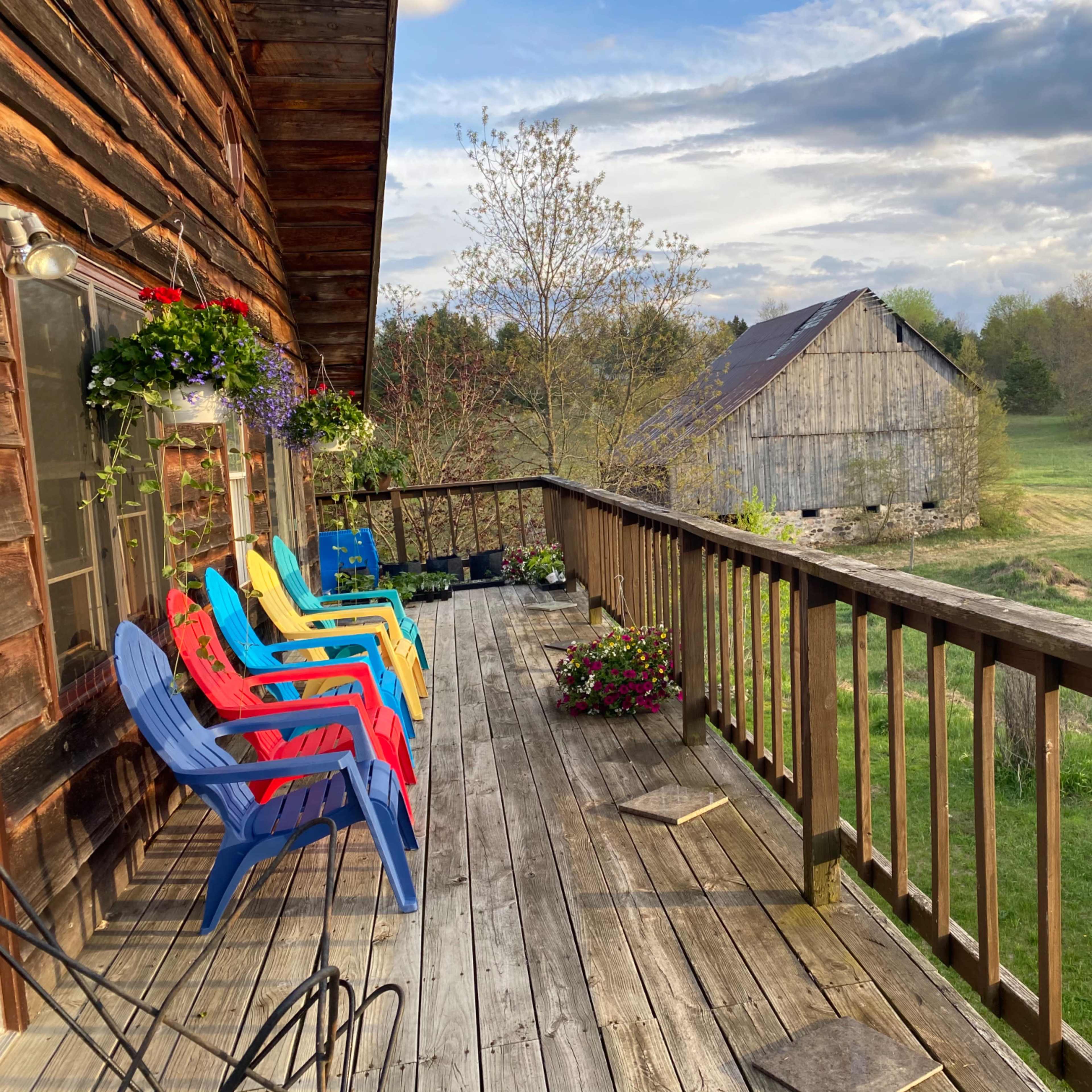 A wooden deck with colorful chairs overlooks a green field and a rustic barn in the distance.