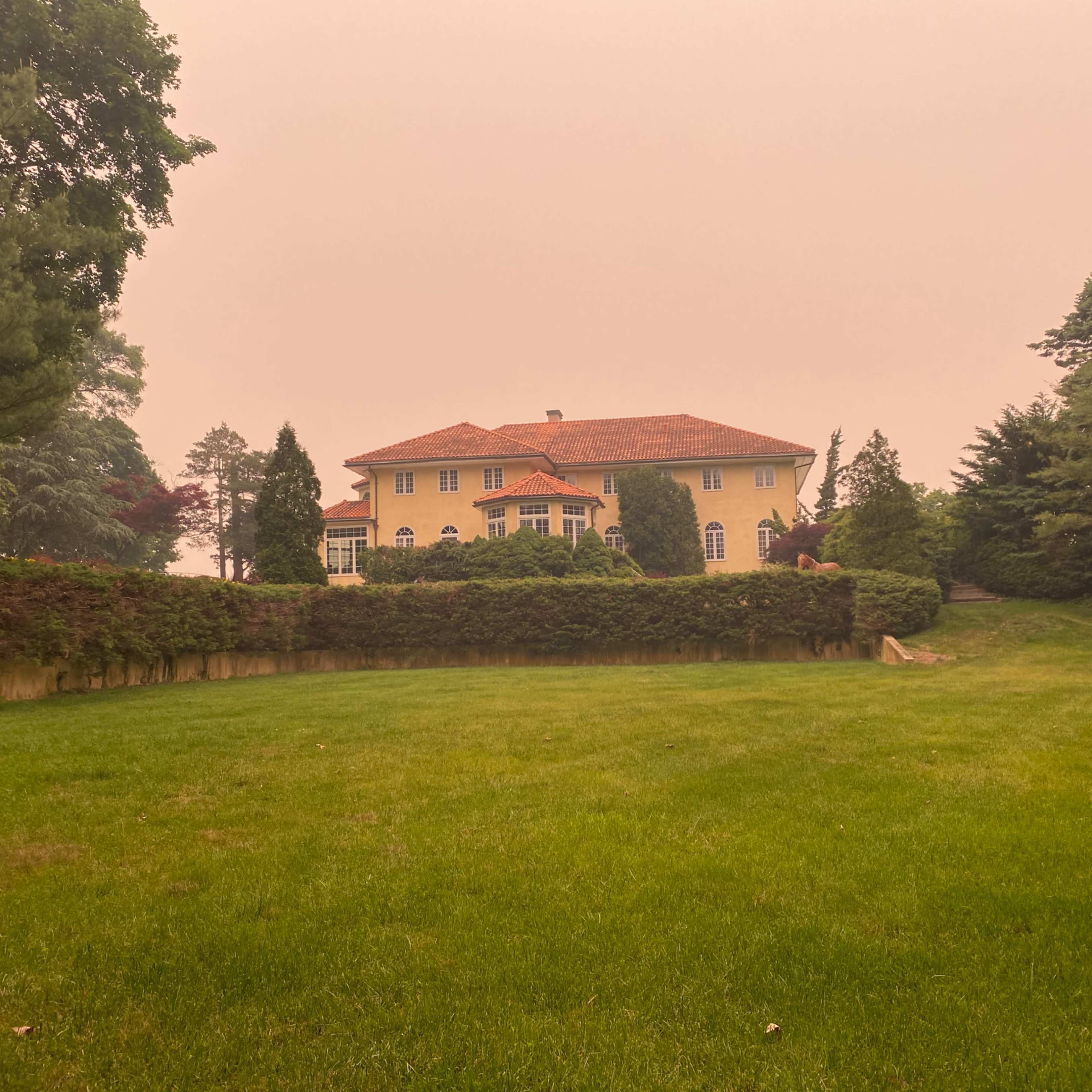 A large yellow mansion with a red tiled roof sits behind neatly trimmed hedges on a green lawn, under a hazy sky.
