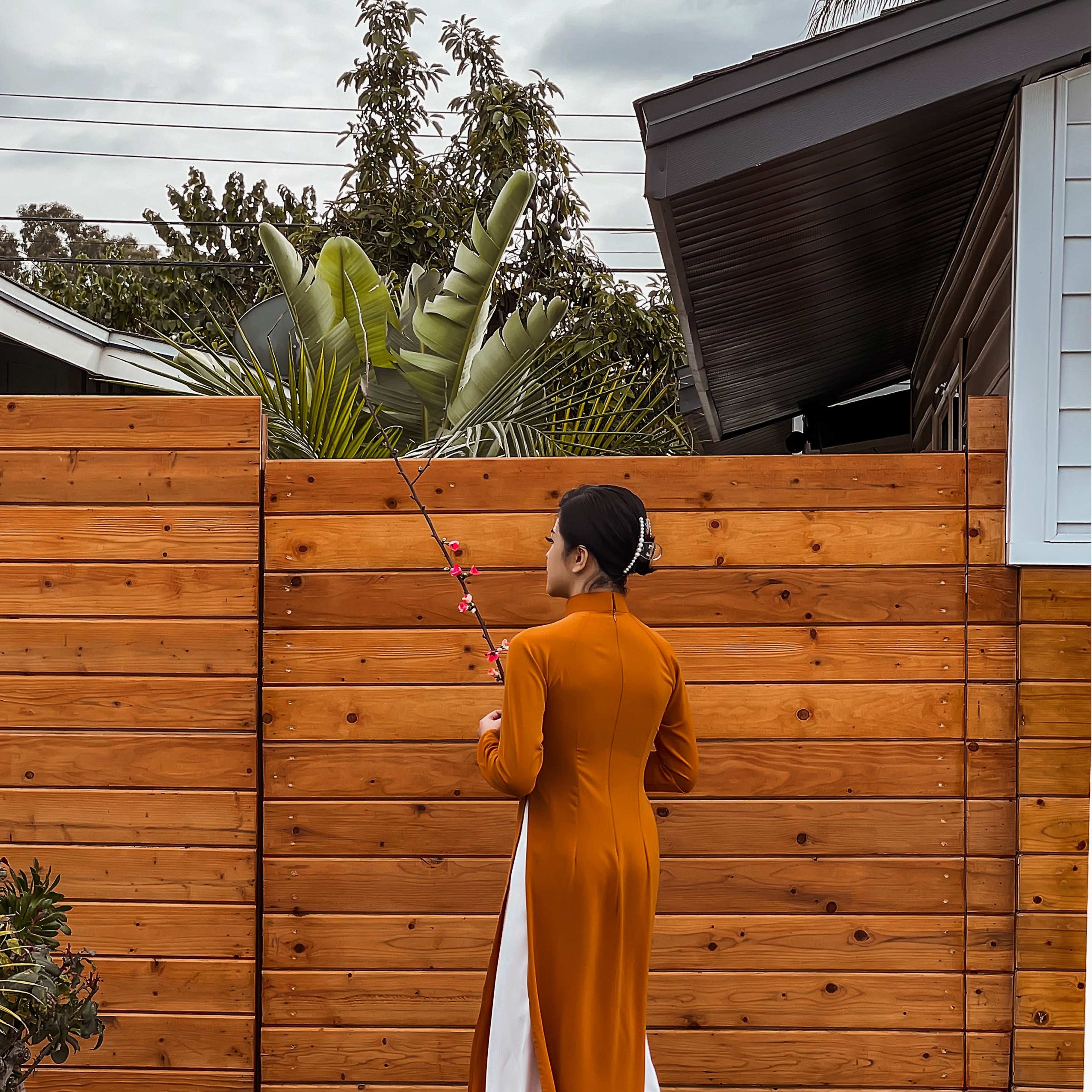 A woman stands facing a wooden fence, wearing a long, brown and white traditional dress, holding a fishing rod near tropical plants.