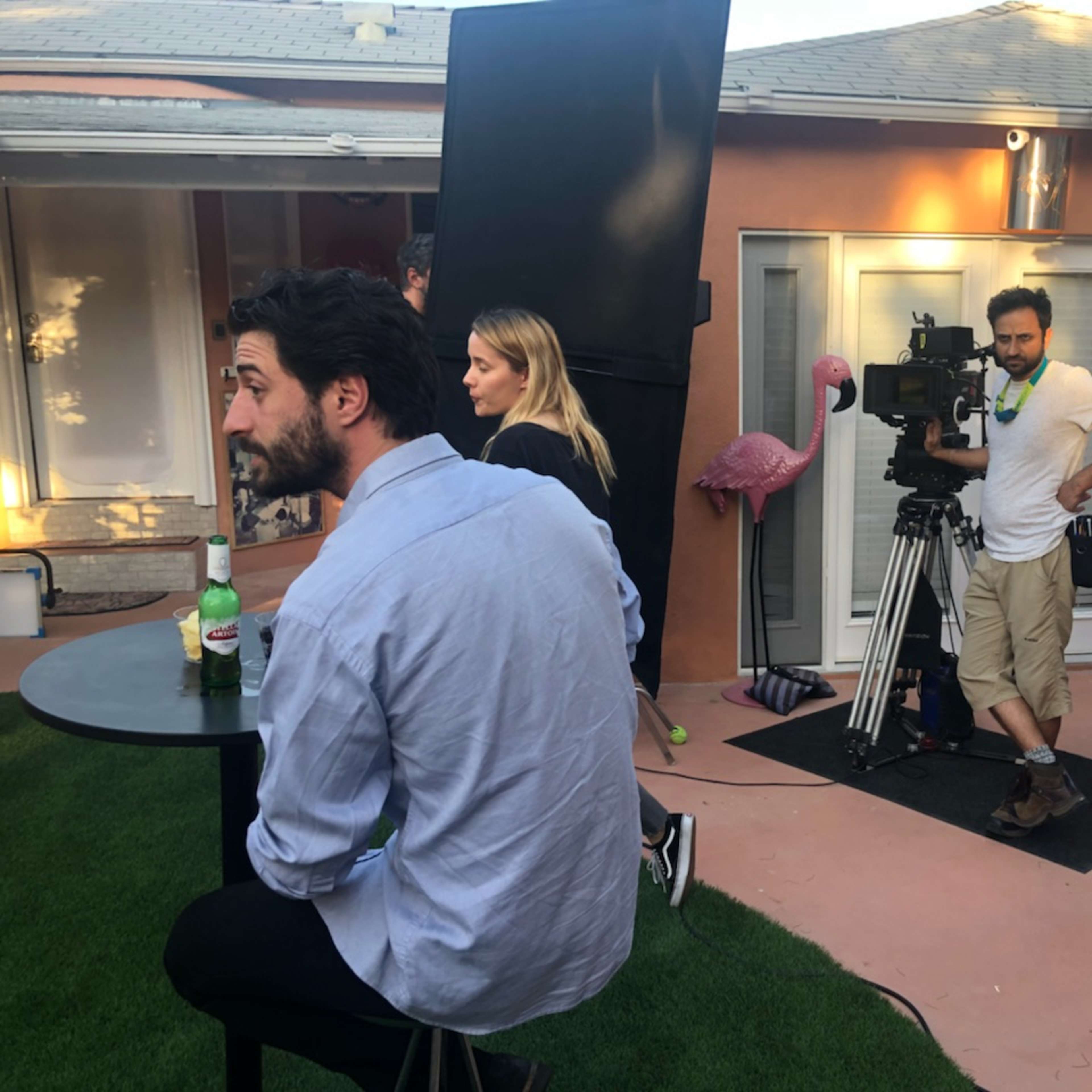 A film crew sets up a scene in an outdoor area with a man seated at a table and additional crew members observing and operating equipment.