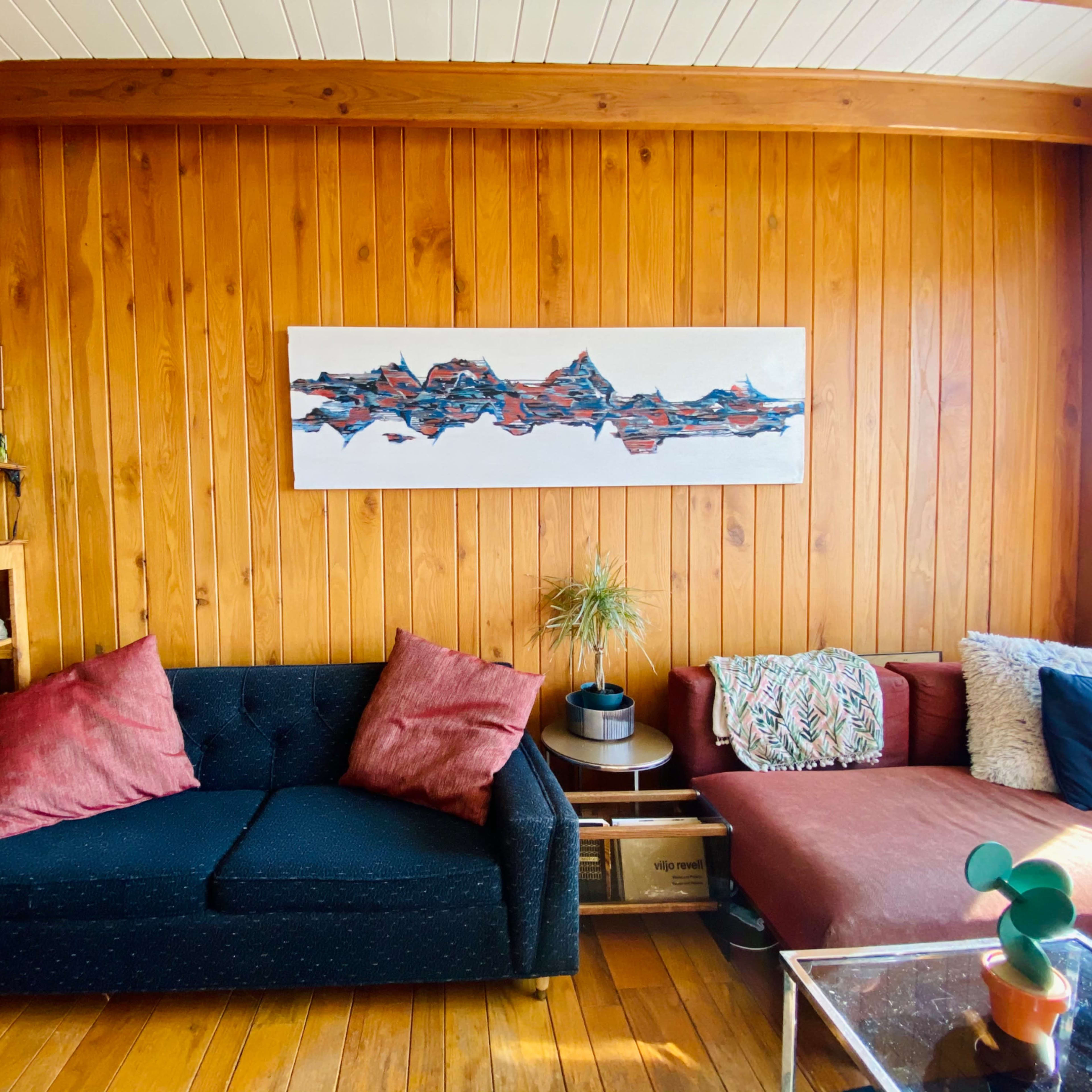 A room with wooden walls featuring two sofas, one dark with red pillows and another in a light color, along with a shelf of items and a large artwork on the wall.