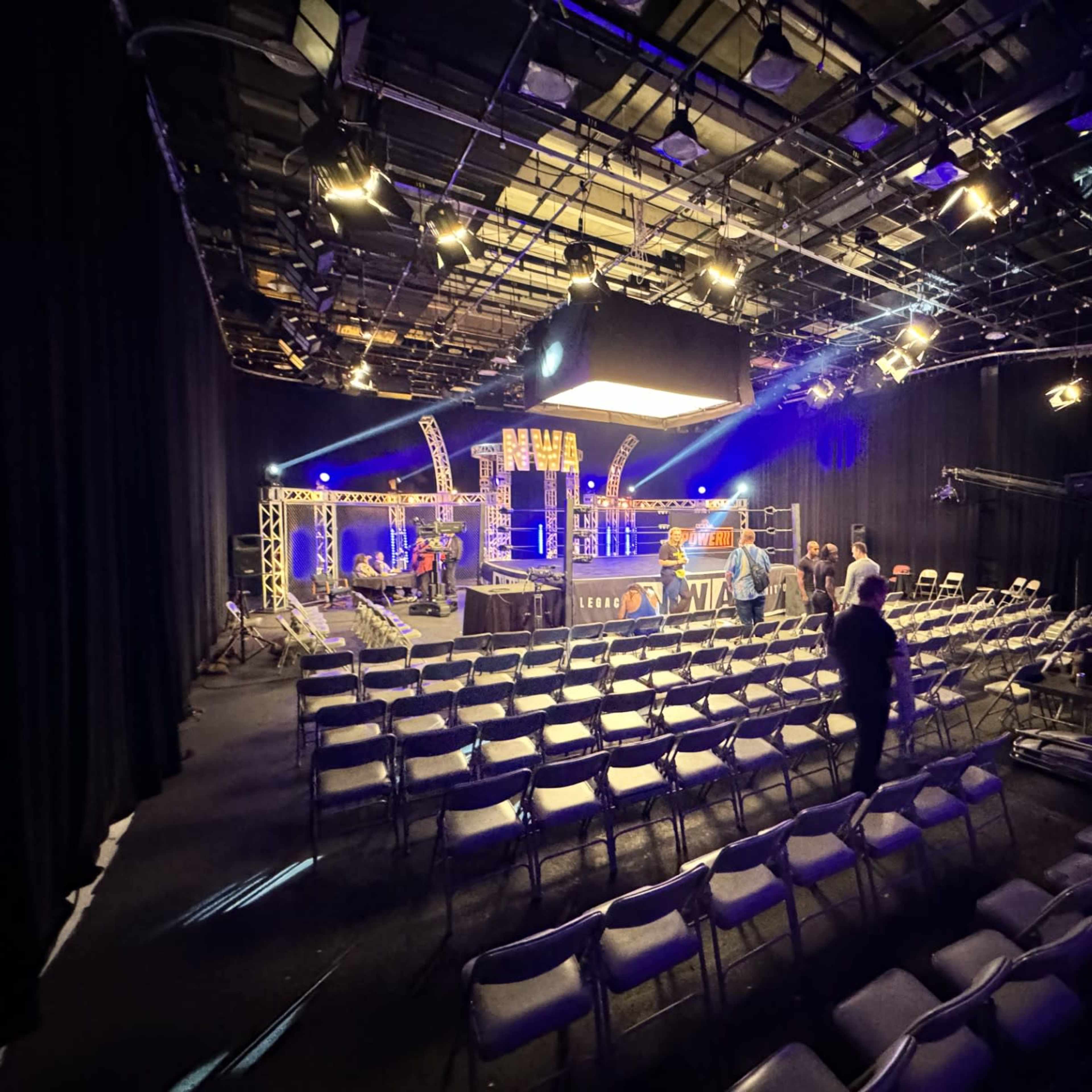 A studio setting with rows of empty chairs facing a well-lit stage, equipped with lighting and camera gear.