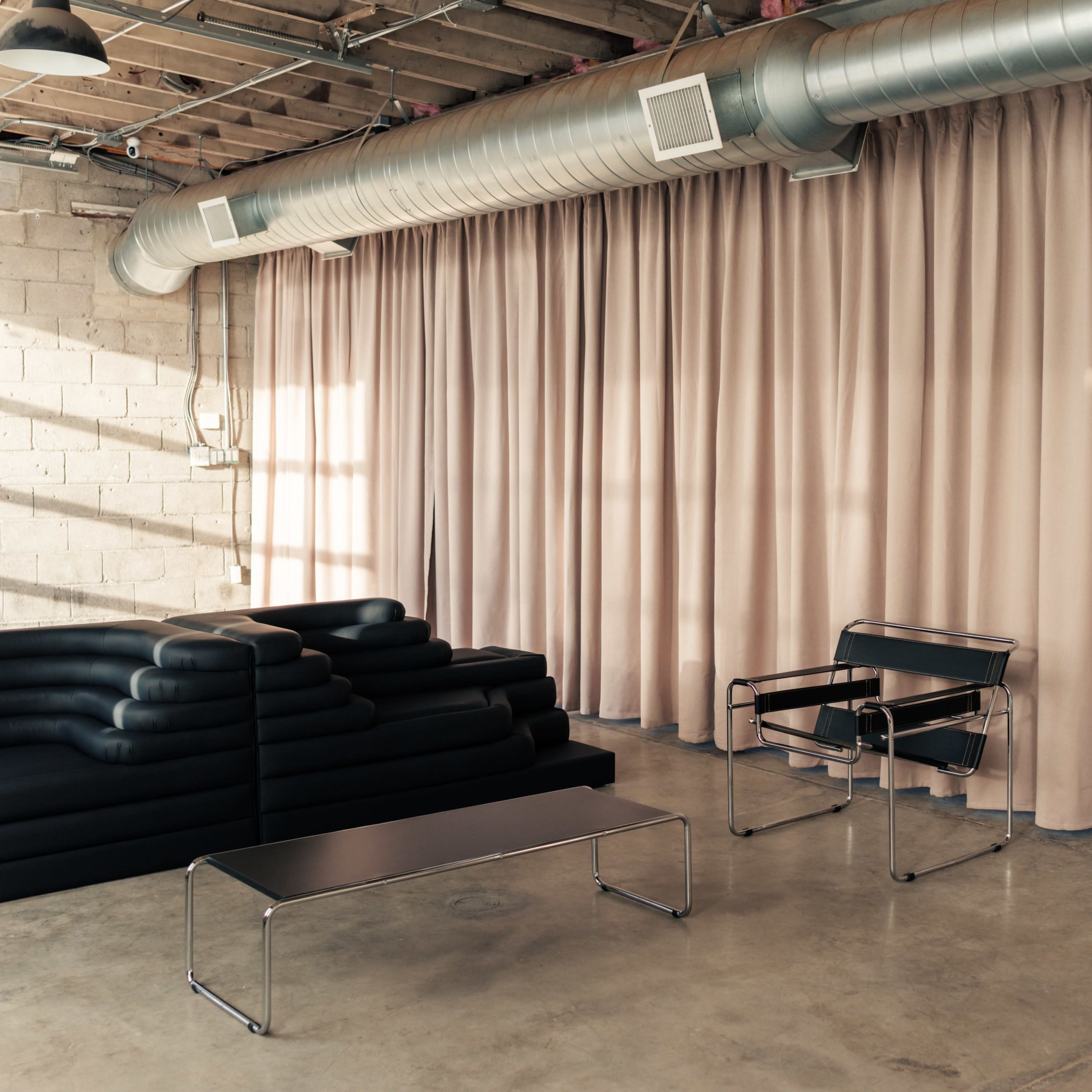 A minimalistic interior featuring a black lounge chair and a low coffee table in front of a wall of beige curtains.