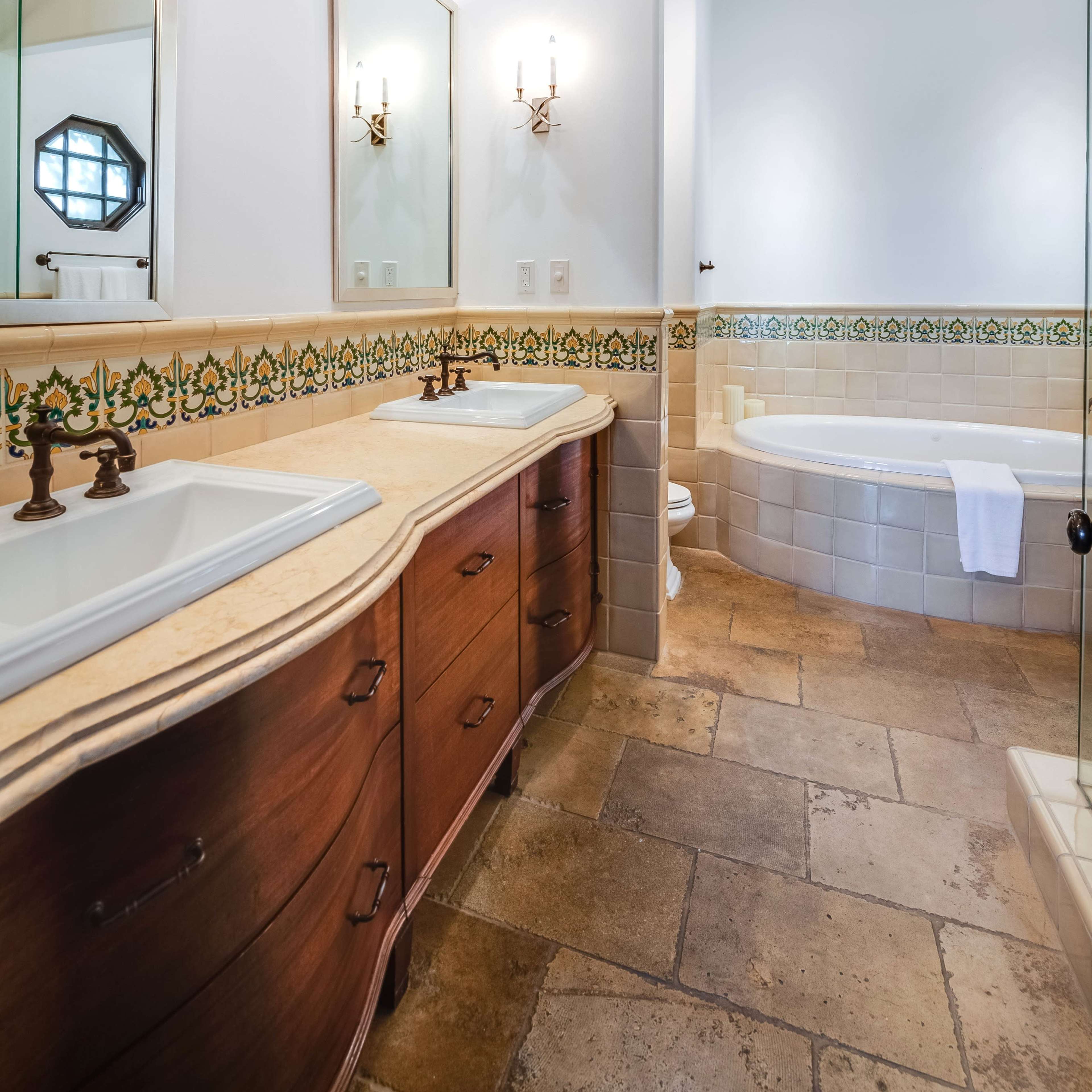 A bathroom featuring a wooden vanity with a dual sink, two mirrors, and a freestanding tub next to tiled walls.