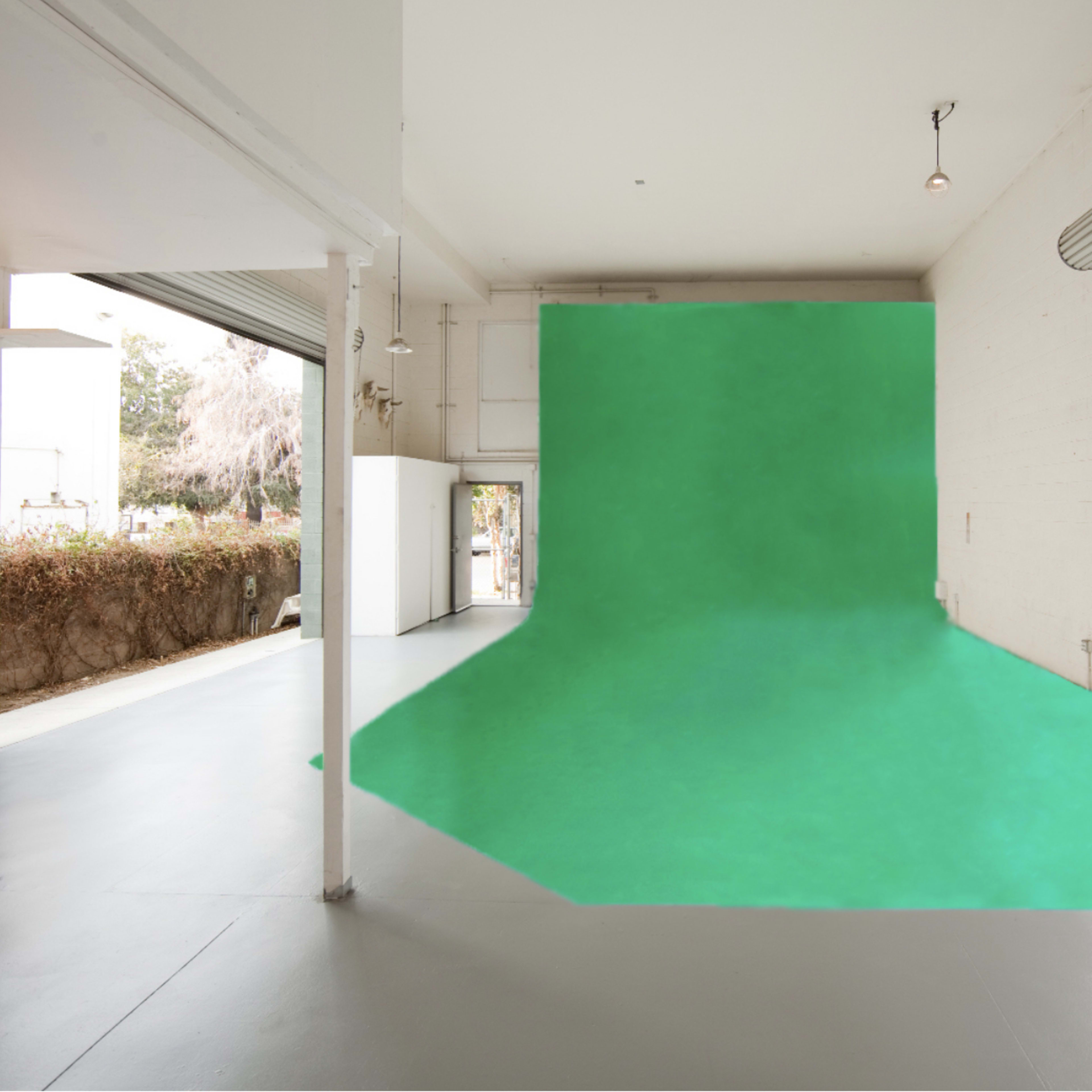 An empty studio space with a green backdrop extending along one wall and floor, surrounded by large windows and a view of outdoor greenery.
