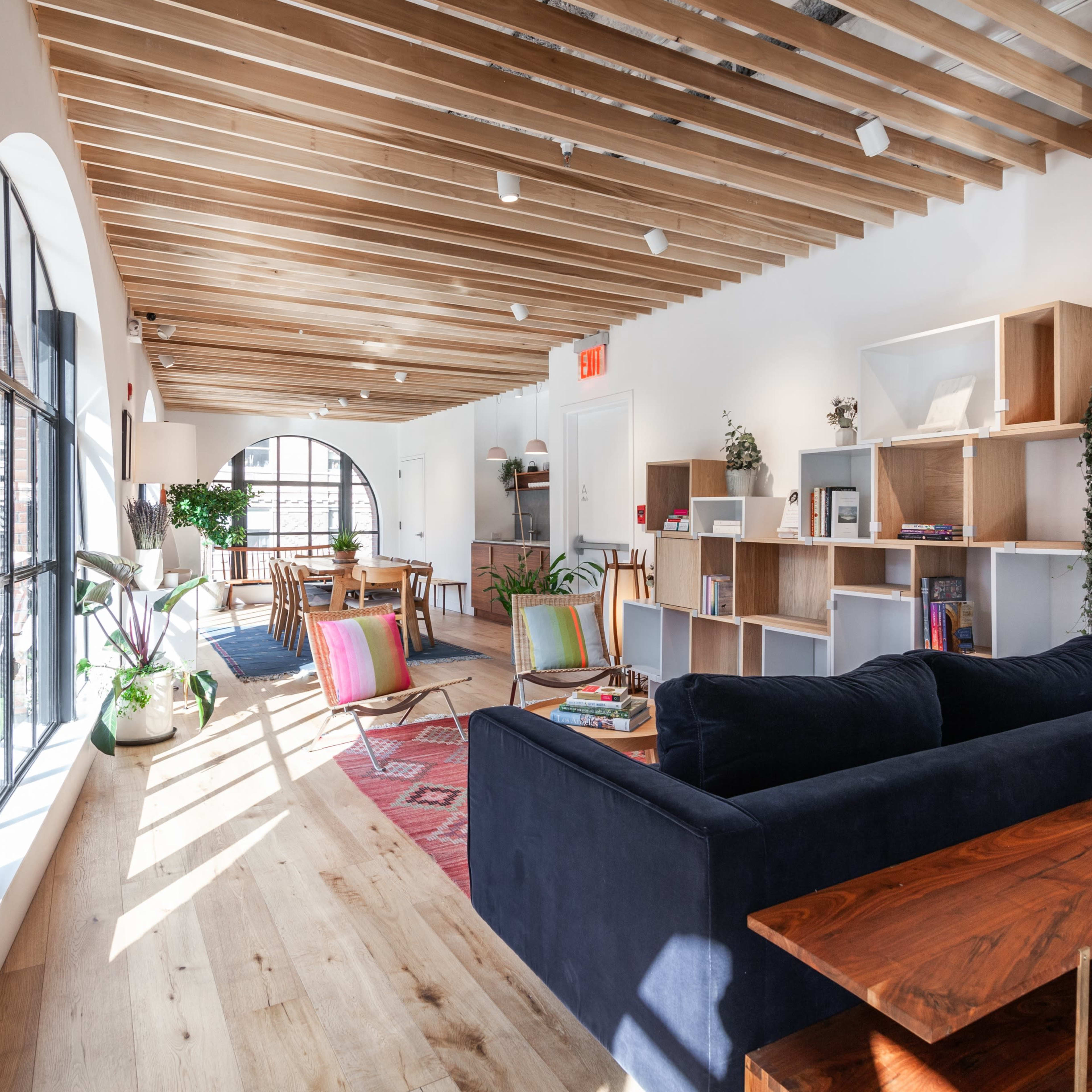 A bright and spacious interior with wooden ceiling beams, large windows, a dark blue sofa, a wooden table, and shelving filled with books and decorative items.