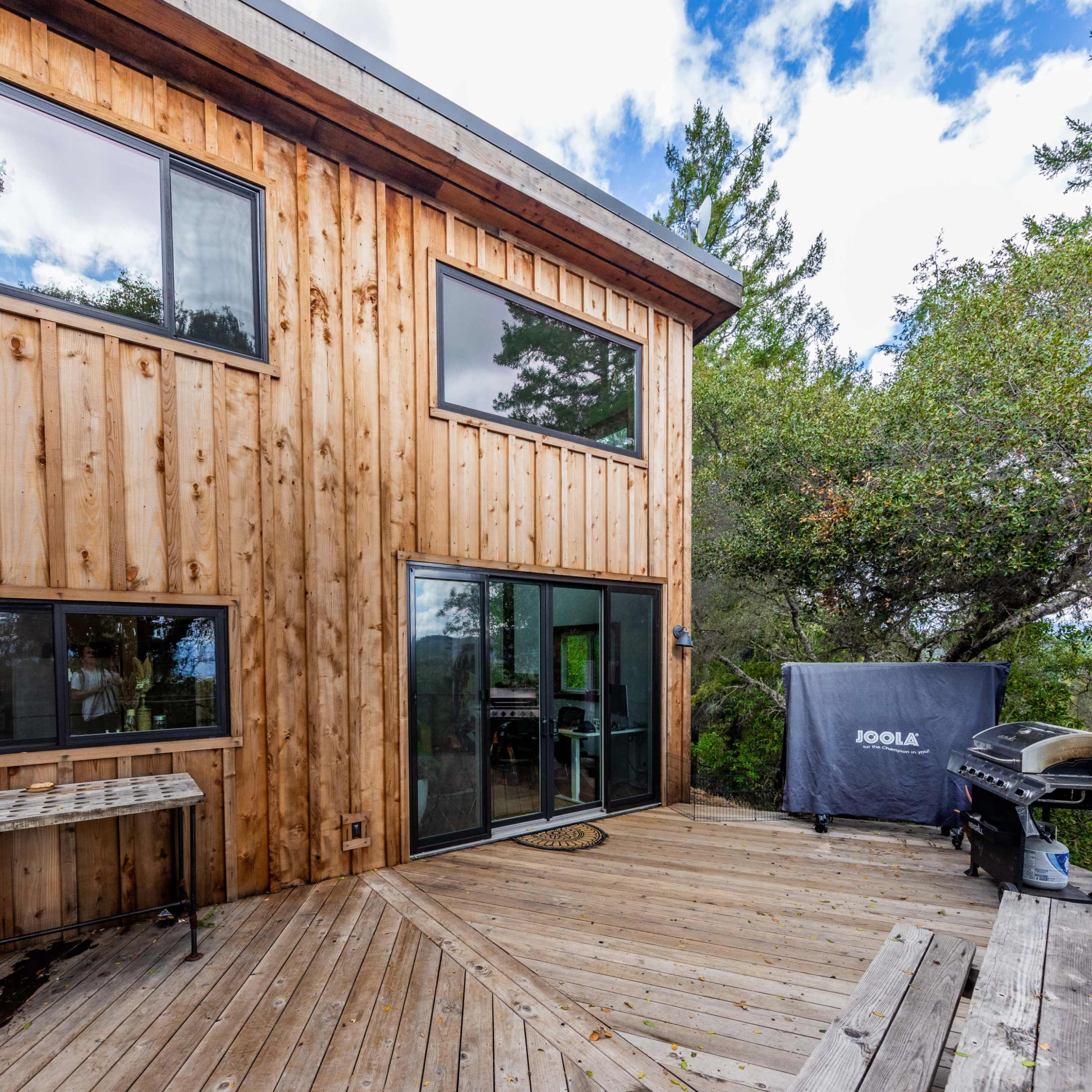 A wooden house with large windows and a deck overlooks a forested area.