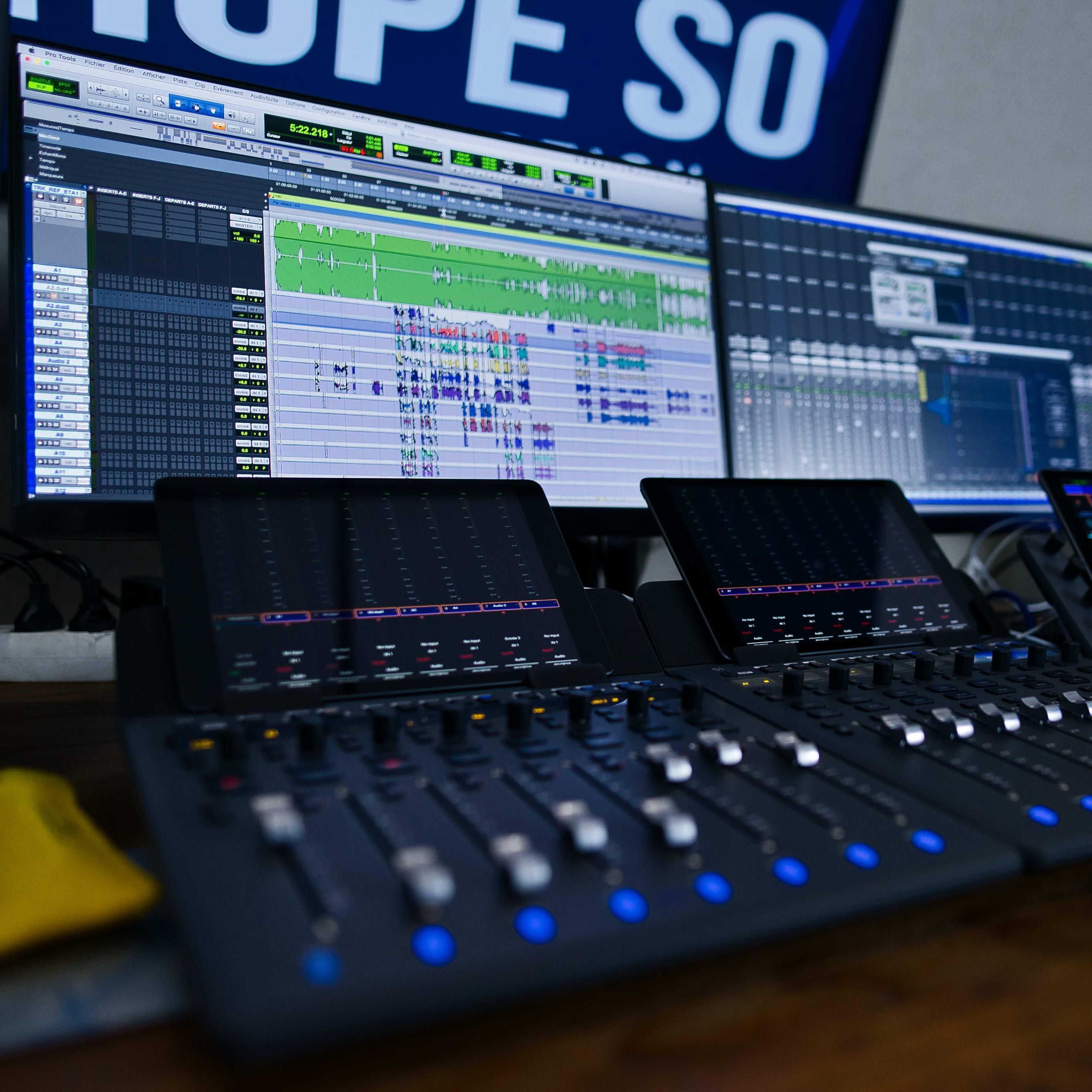 A digital audio workstation with two large monitors displaying audio editing software, a control surface with sliders and buttons, and studio speakers on either side.