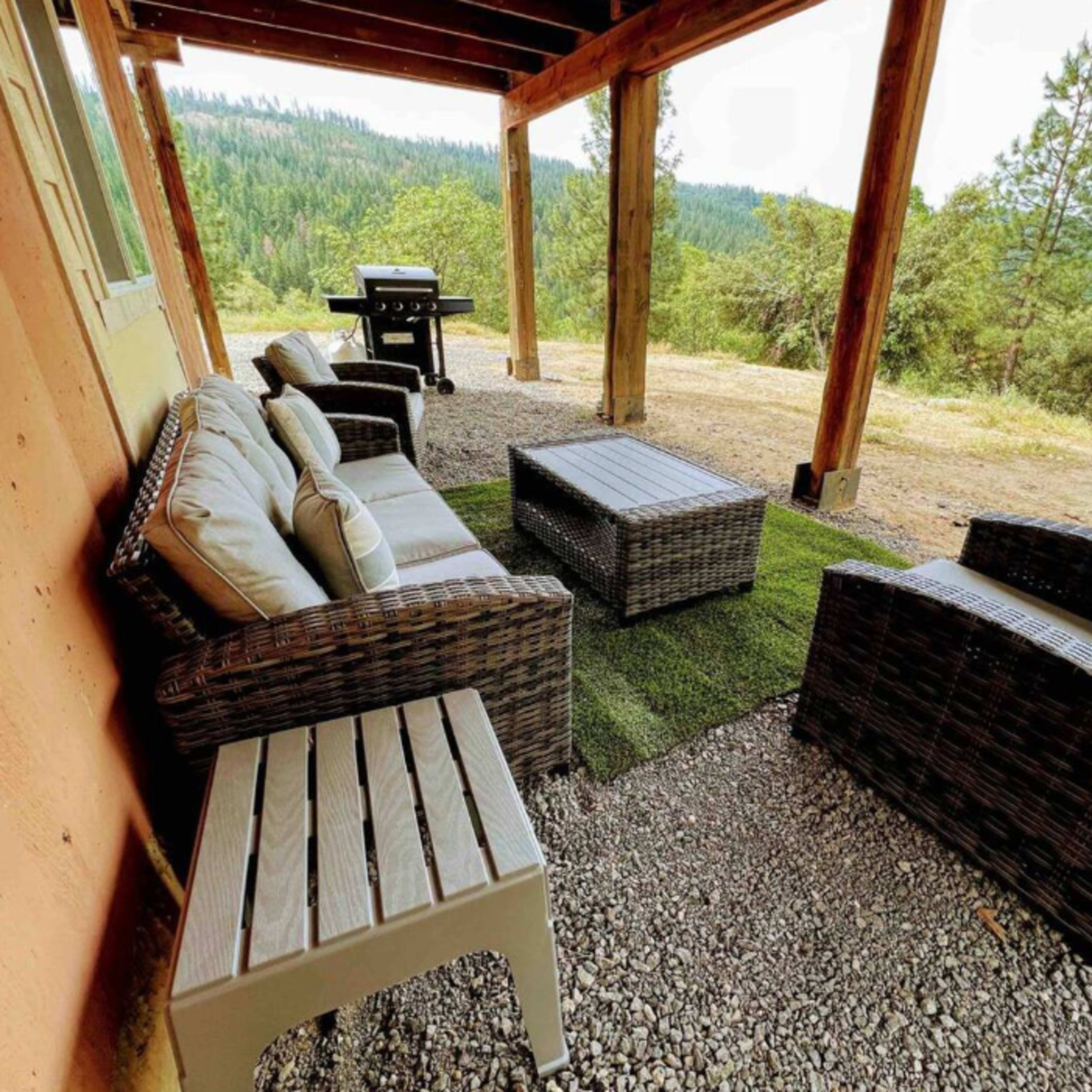 A covered outdoor seating area features a couch, two chairs, and a coffee table on a grassy patch, with a grill nearby and a view of trees in the distance.