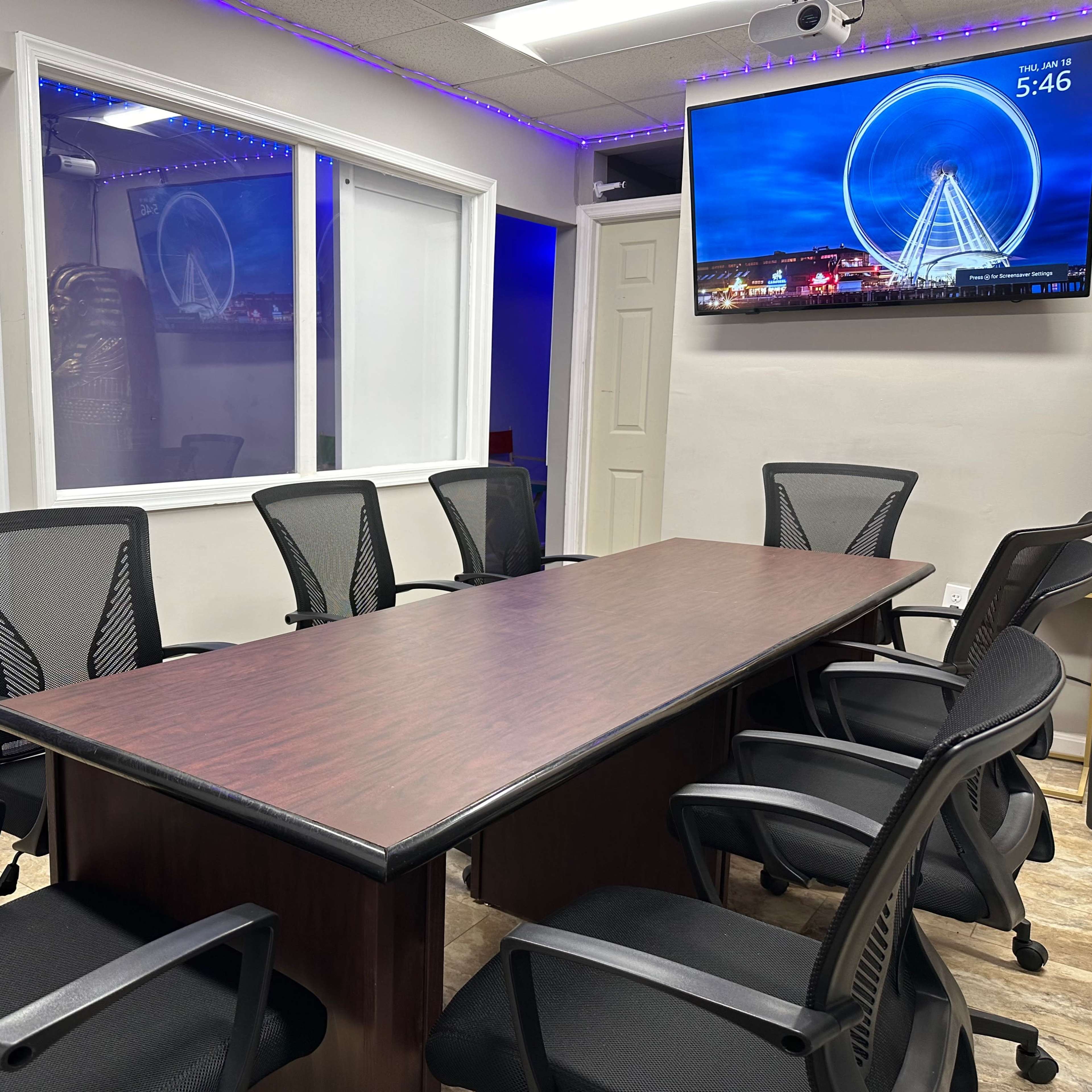 A conference room features a large wooden table surrounded by six black mesh chairs, with a television displaying a Ferris wheel image on the wall.