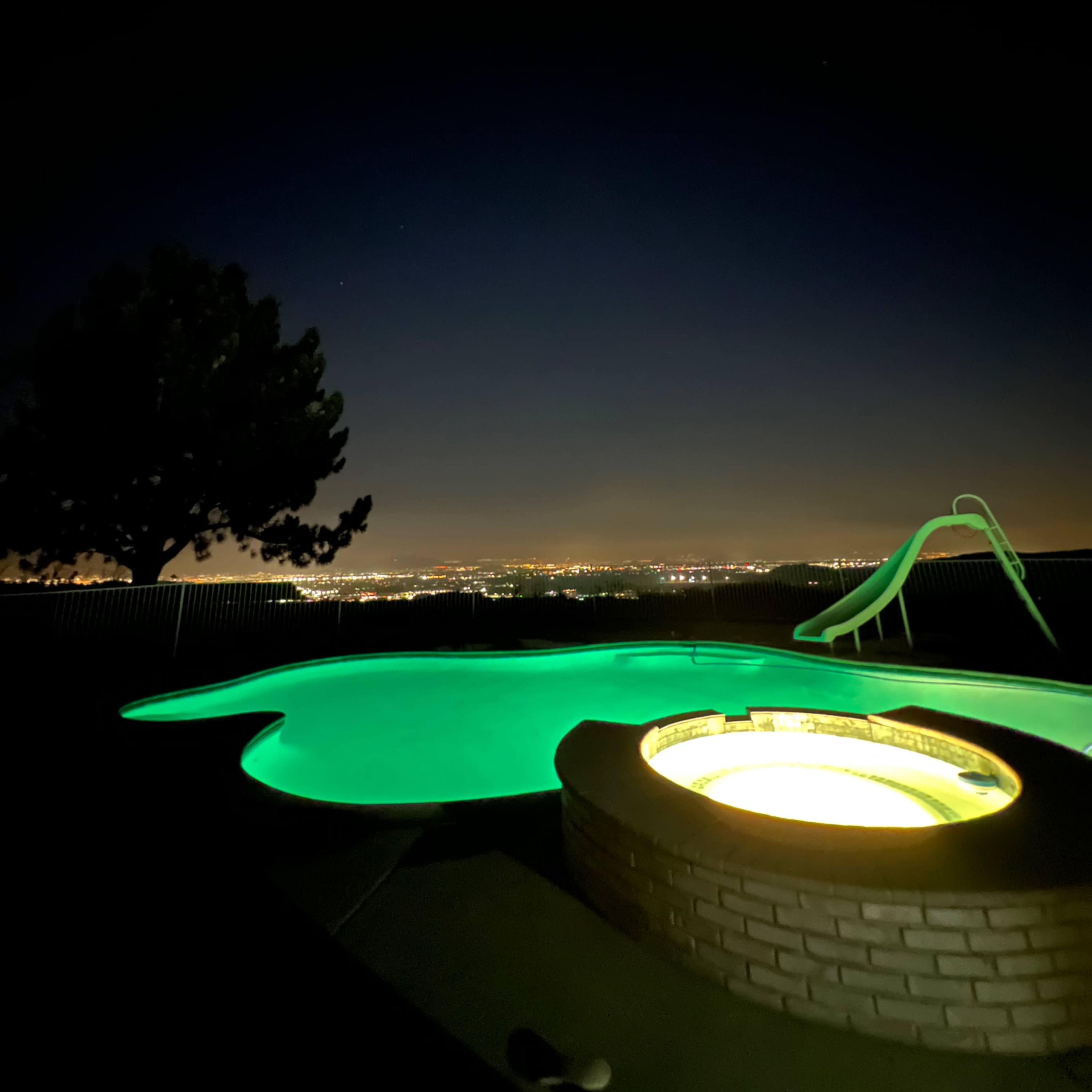 A brightly lit hot tub beside a dark swimming pool, set against a city skyline at night.