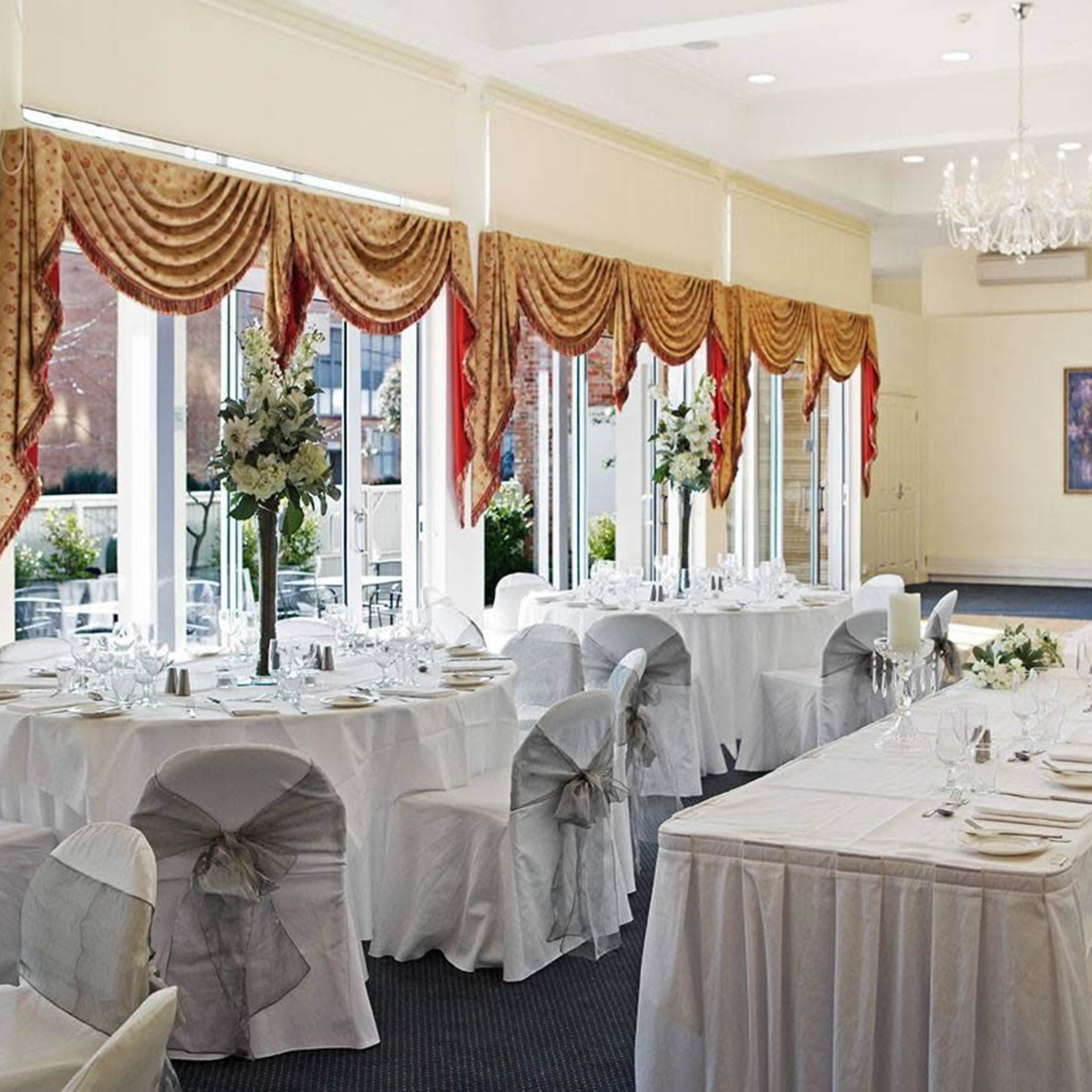 A banquet room is set up for an event with neatly arranged tables covered in white linens and decorated with floral centerpieces near large windows.
