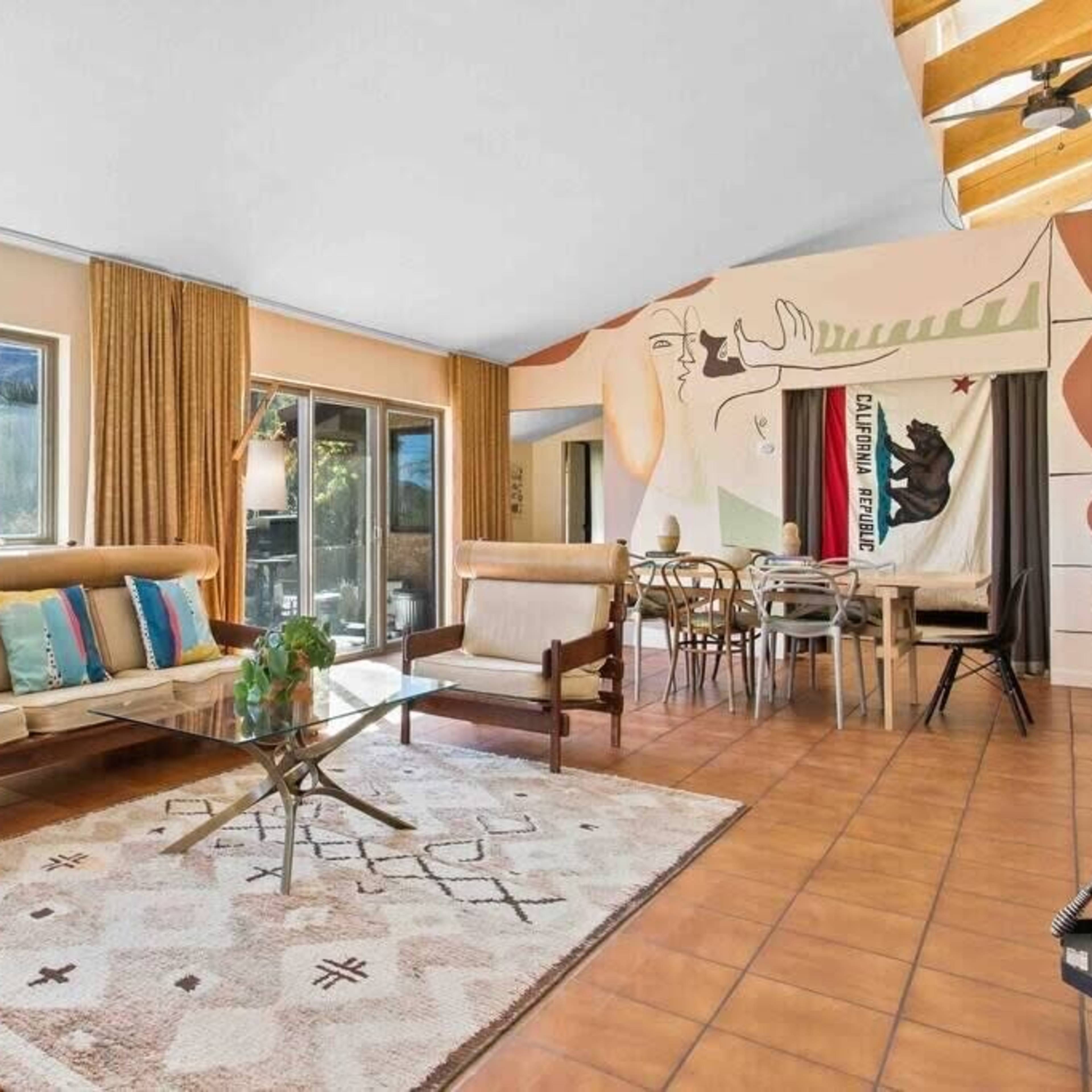 The living room features a beige sofa with colorful pillows, a glass coffee table, and large windows illuminating a mural-adorned wall.