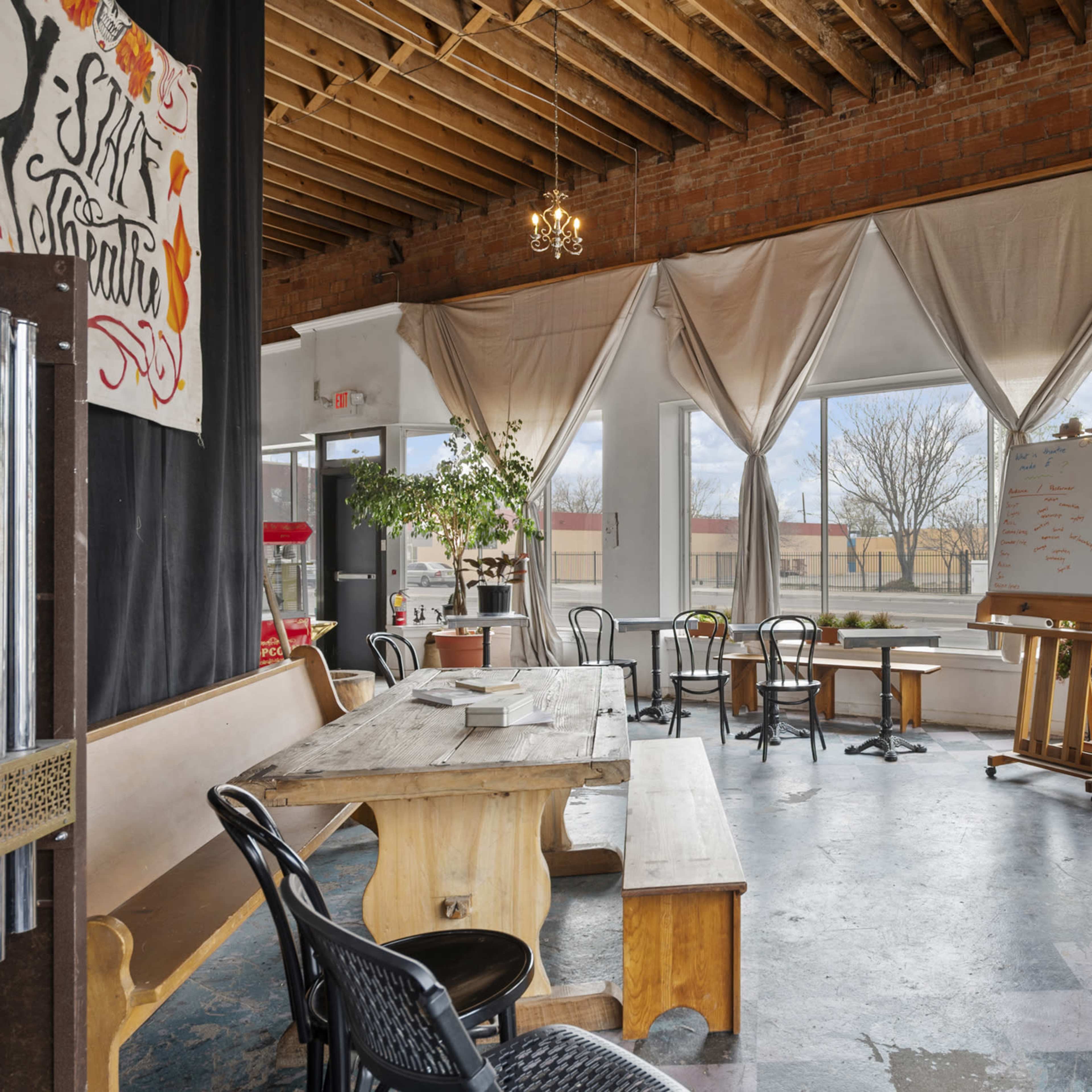 A spacious café interior with a mix of wooden tables and chairs, large windows allowing natural light, and a whiteboard at the back.