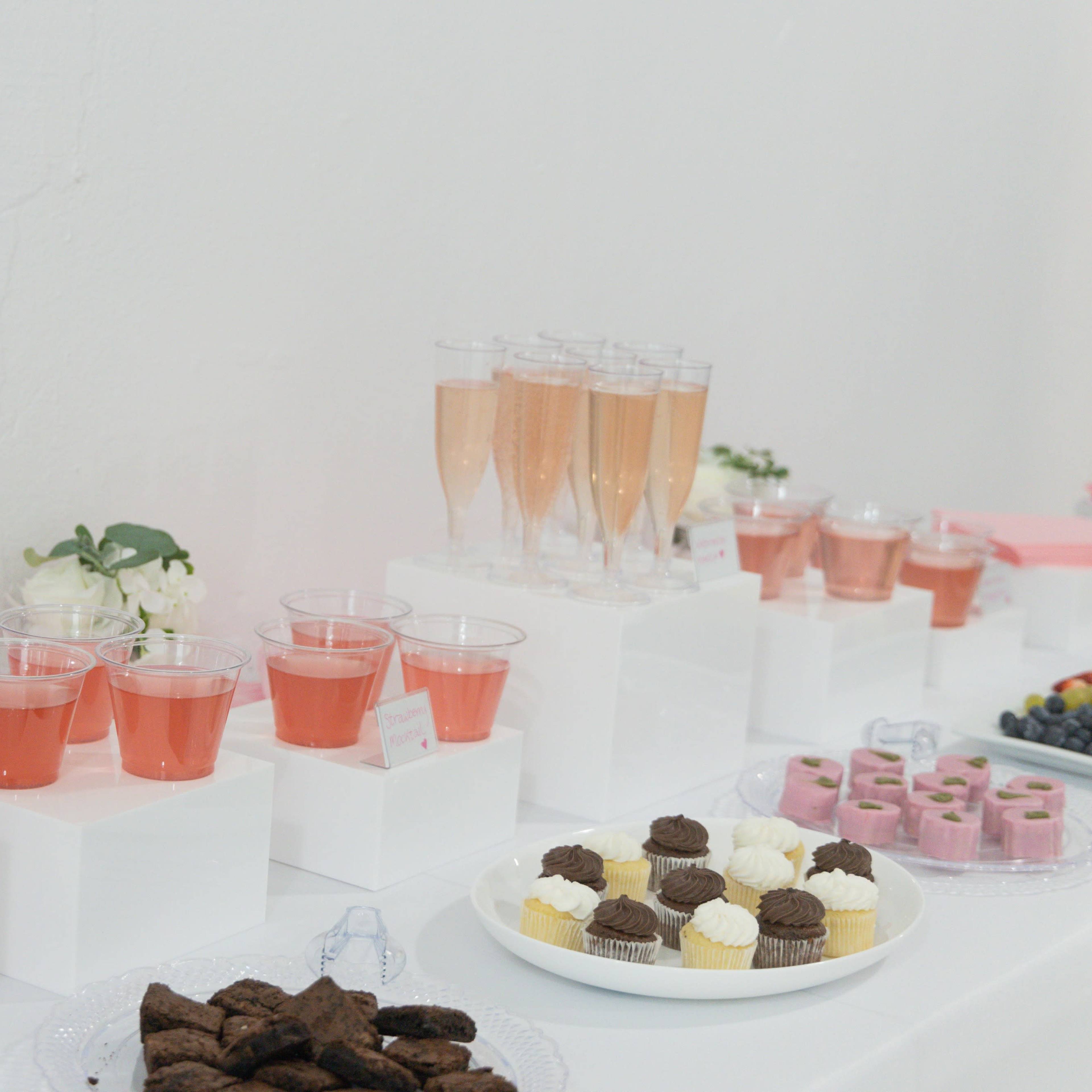 A table displaying various drinks in clear glasses, including pink beverages, alongside an assortment of desserts such as cupcakes, cookies, and gelatin treats.