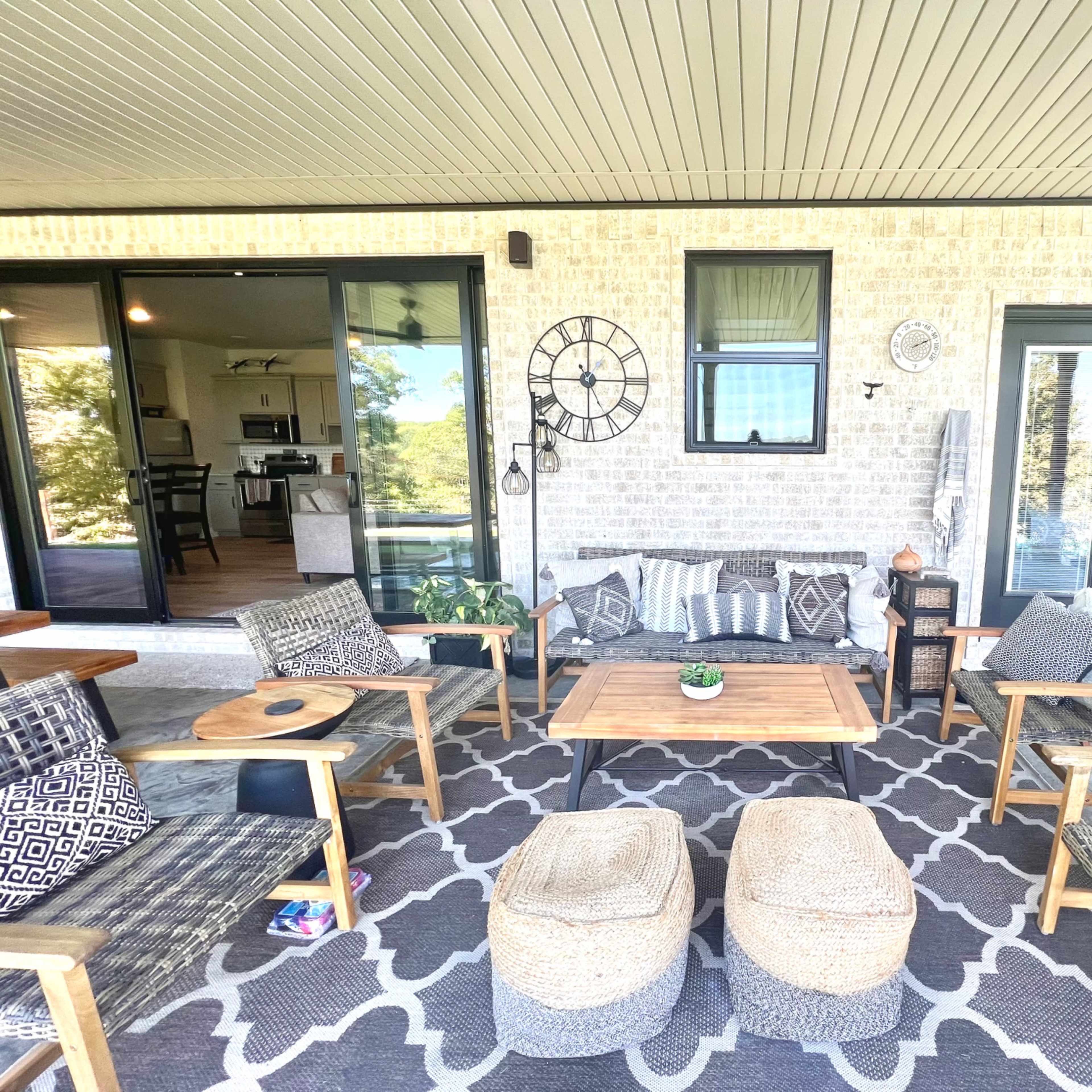 A covered outdoor seating area features a wooden table surrounded by woven chairs and poufs, with a patterned rug and wall decorations.