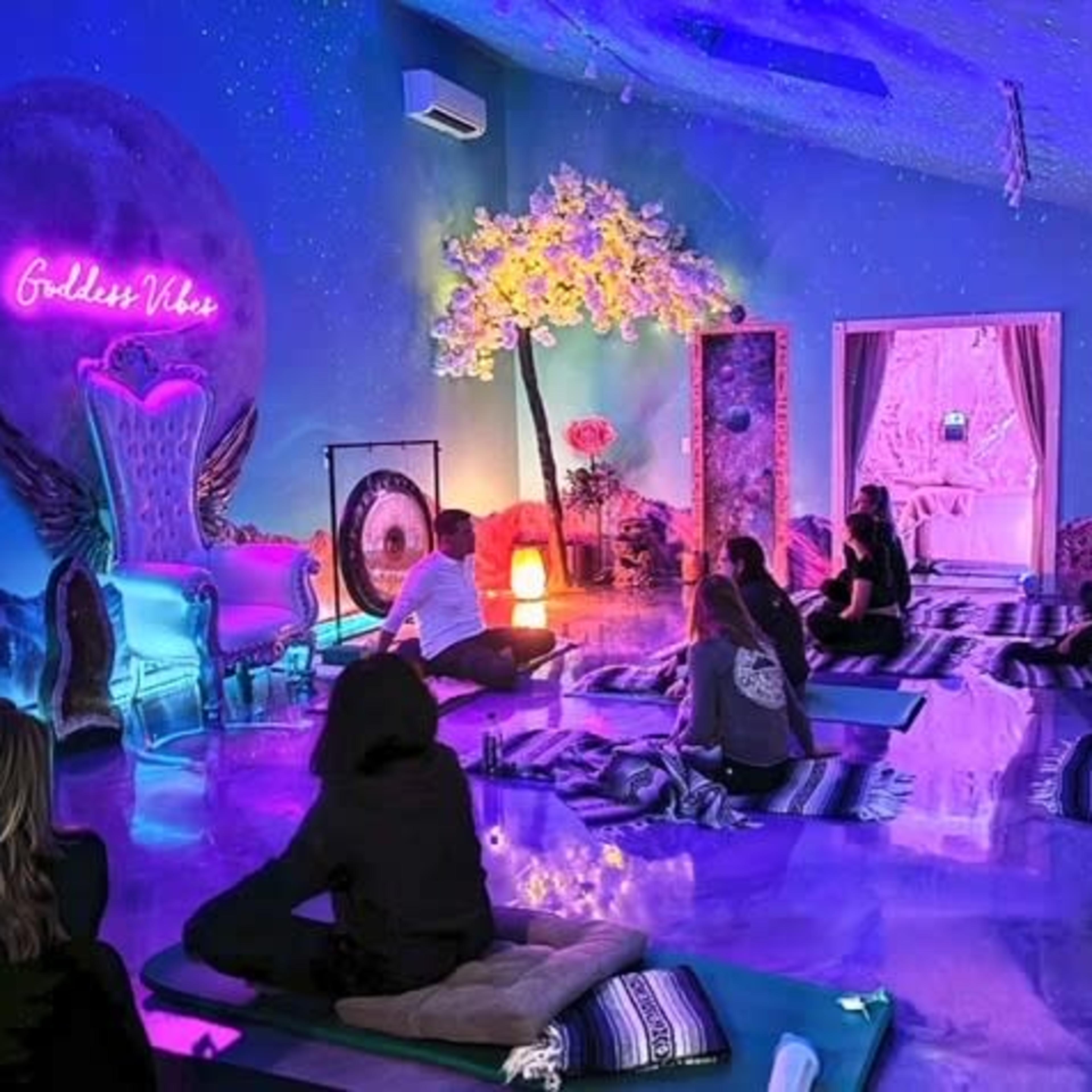 A group of people sits on mats in a brightly colored room adorned with neon lights, flowers, and a decorative arch.